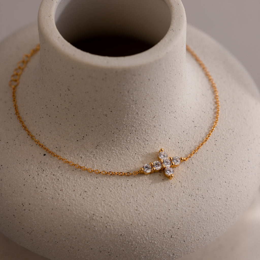 Gold chain bracelet featuring a delicate cross pendant set with round, sparkling clear stones, displayed on a neutral ceramic vase.