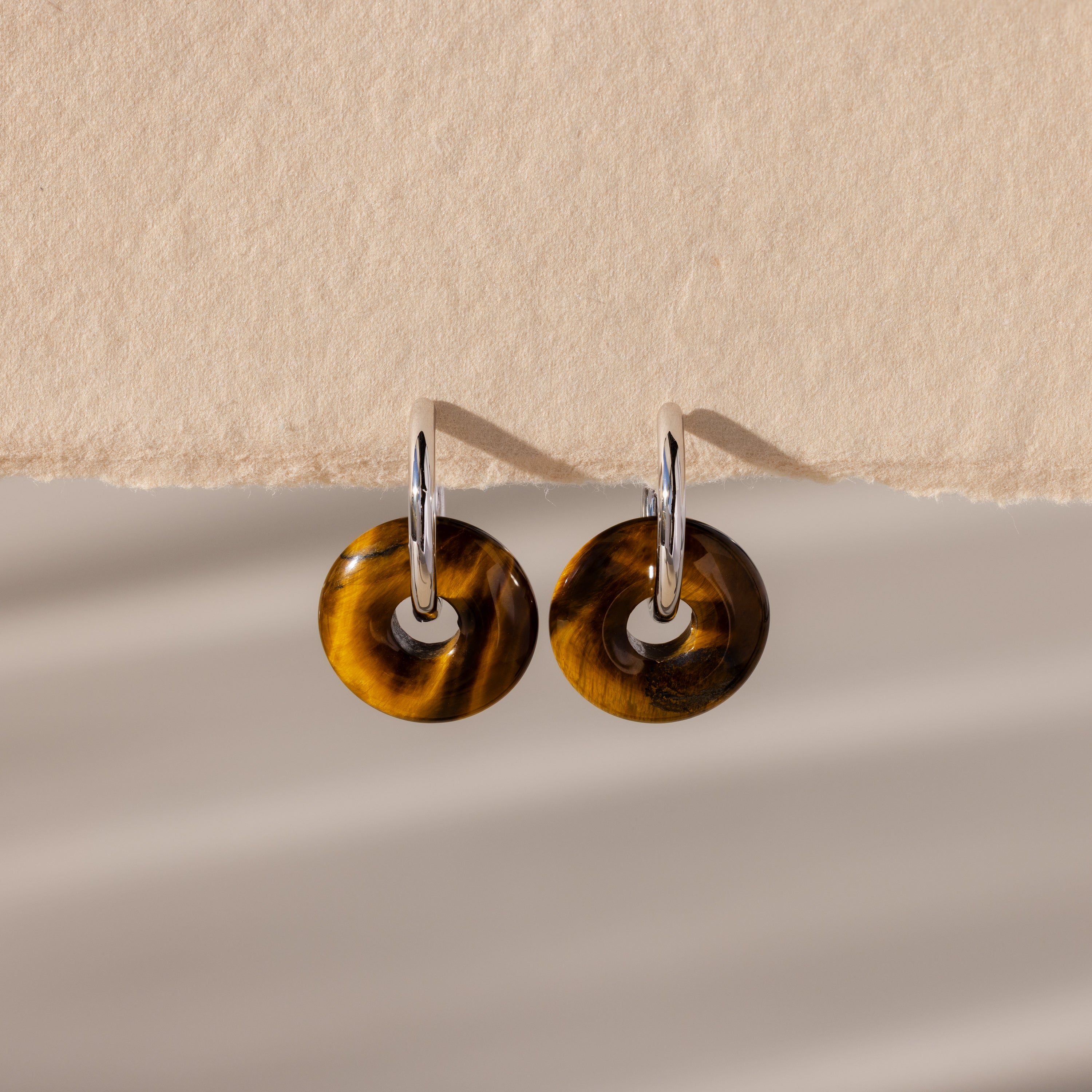 Tiger's Eye Duo Hoops in White Gold, featuring round brown tortoiseshell pendants inspired by golden-brown jewelry, displayed on a textured beige background.