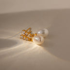 Diamond Pearl Droplet Earrings in 18K Gold, featuring pearls and small crystals, rest on a beige surface illuminated by soft lighting and gentle shadows.