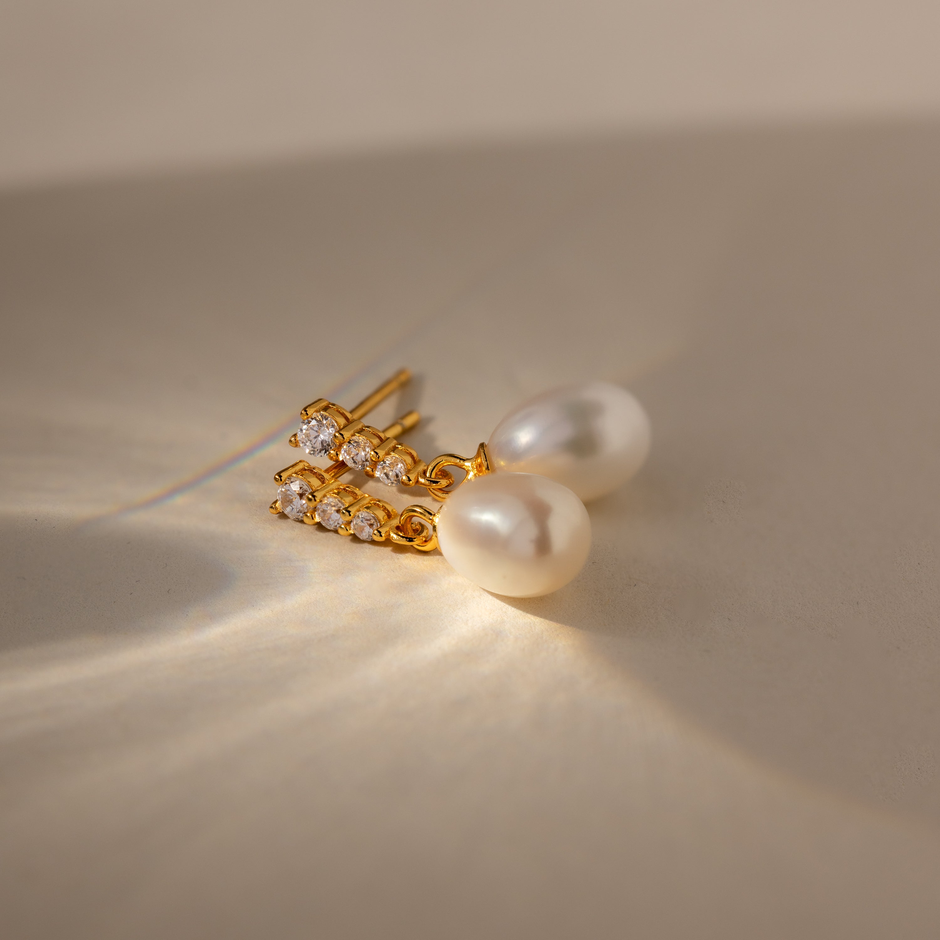 Diamond Pearl Droplet Earrings in 18K Gold, featuring pearls and small crystals, rest on a beige surface illuminated by soft lighting and gentle shadows.
