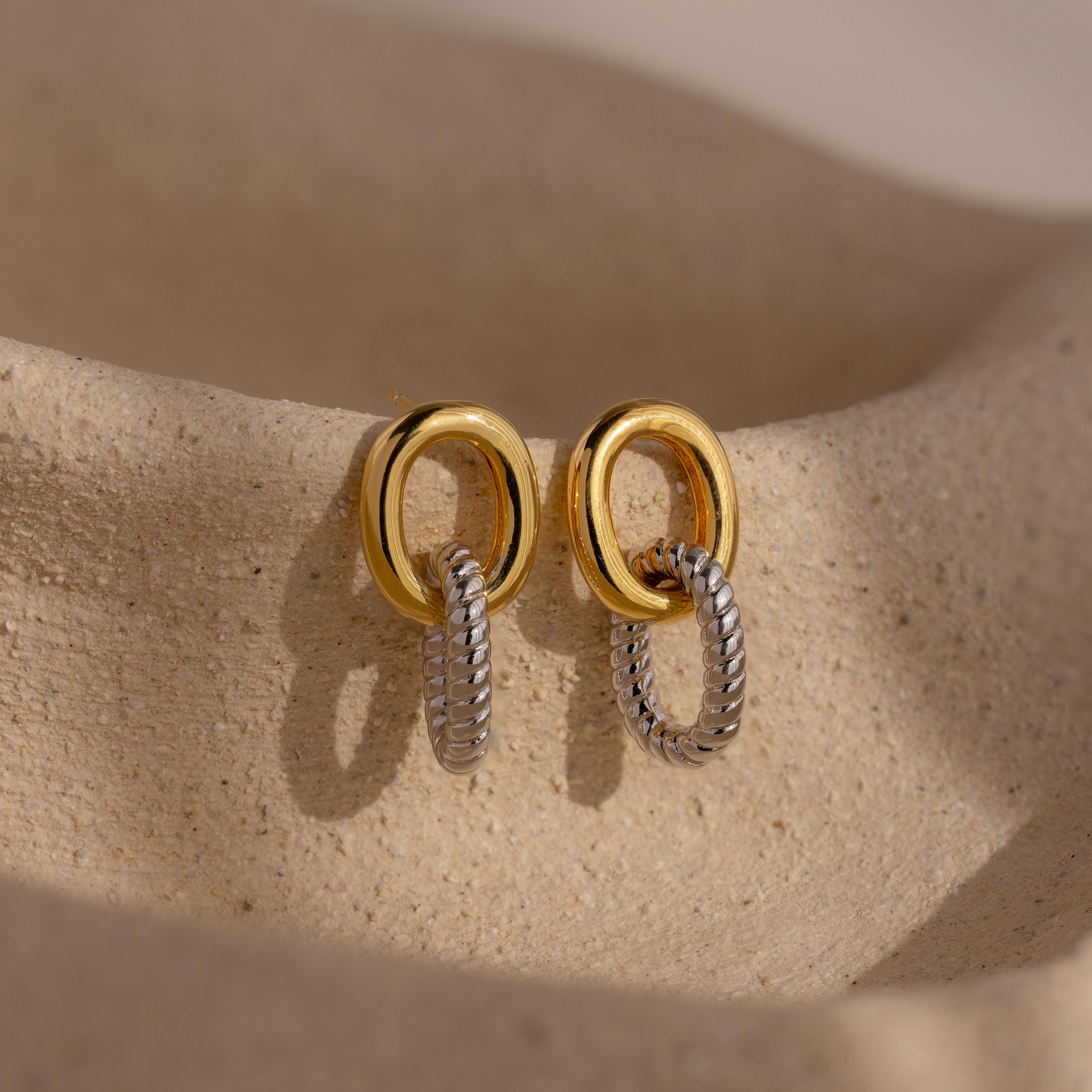 Twisted Link Earrings in gold and silver are displayed on a textured beige surface under soft natural light, highlighting their contemporary design.