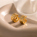 Pave Checkered Huggies, featuring square patterns and embedded crystals, rest on a soft beige dish with sheer fabric in the background.