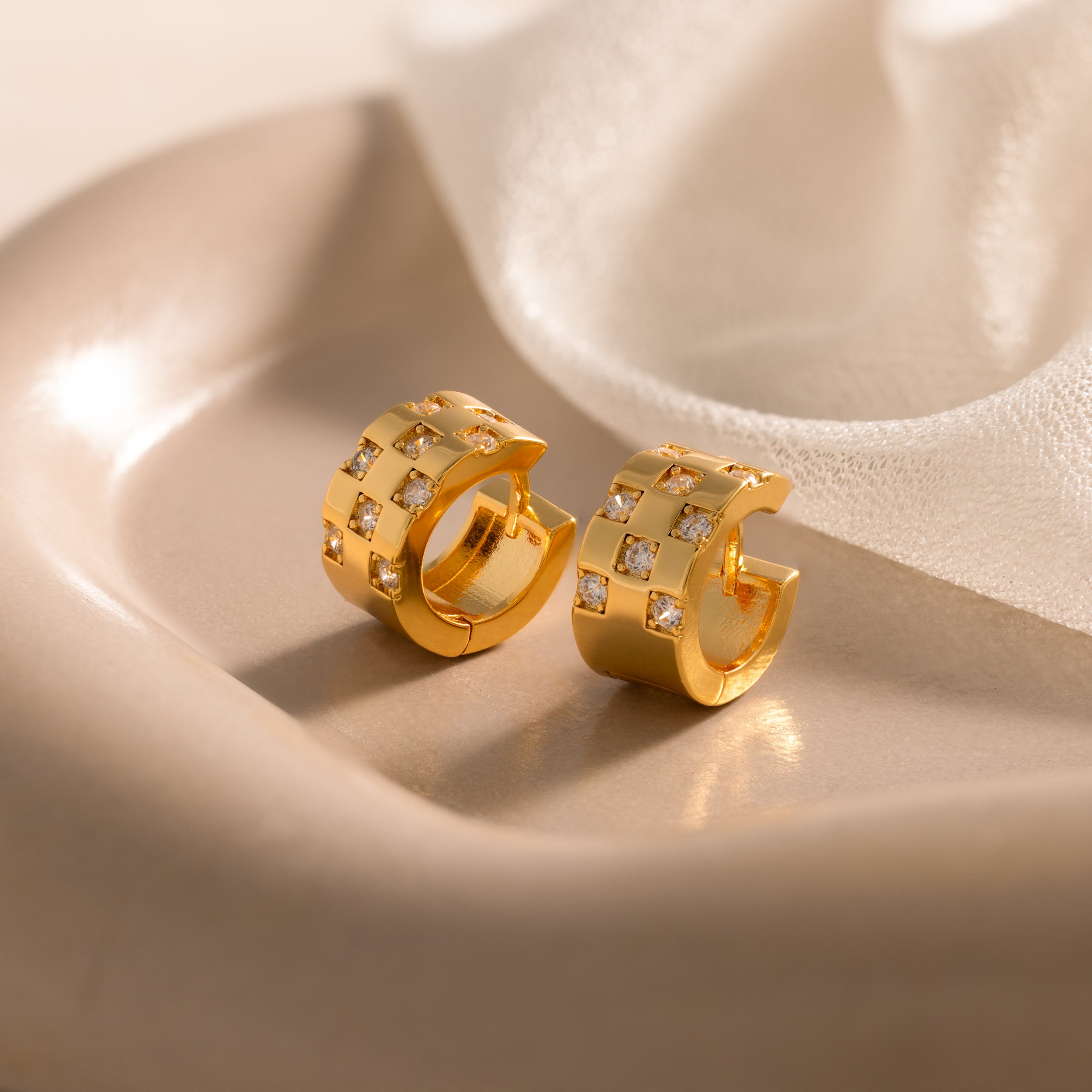 Pave Checkered Huggies, featuring square patterns and embedded crystals, rest on a soft beige dish with sheer fabric in the background.