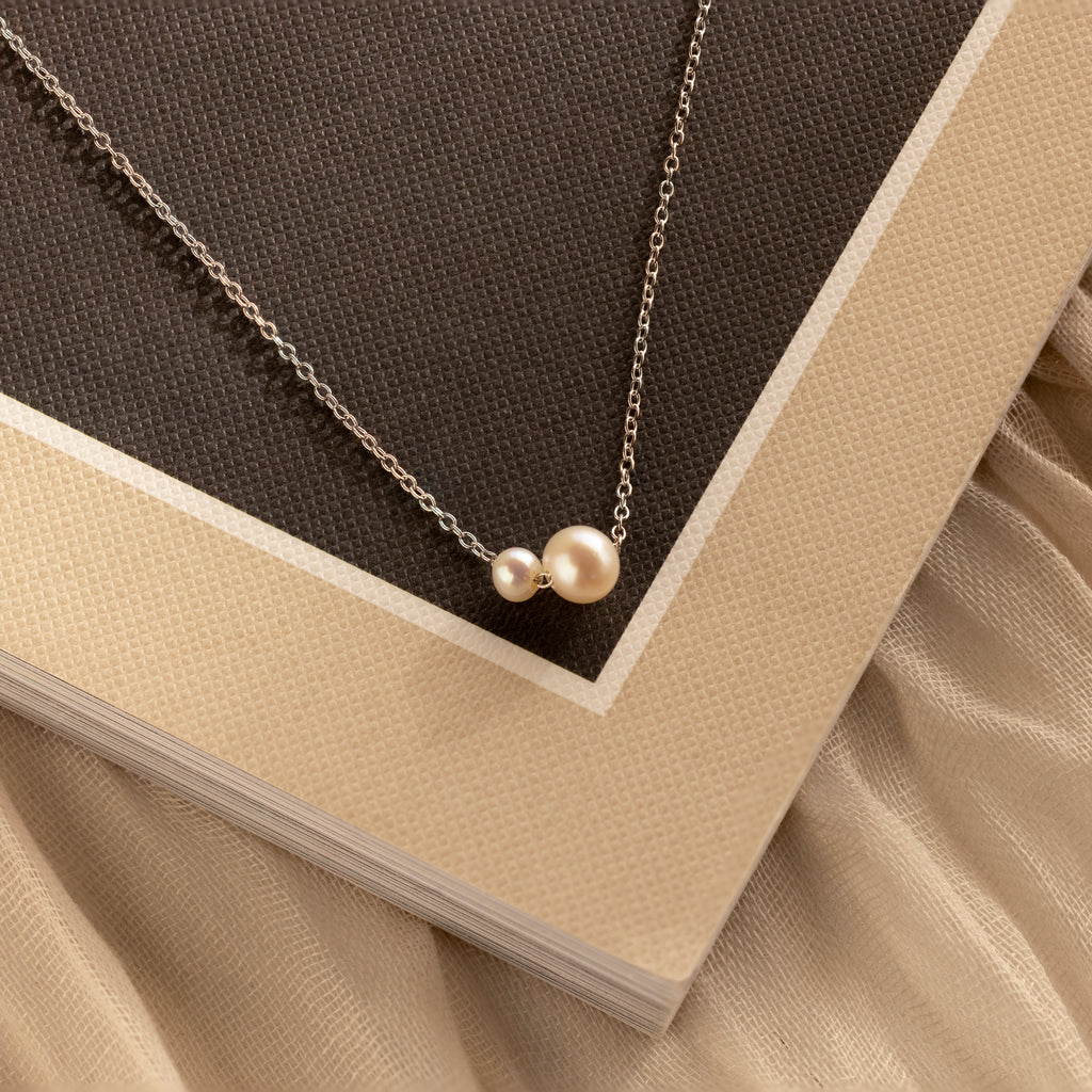 The Dainty Duo Pearl Necklace in Silver features two pearl pendants, shown on a beige and brown book—an elegant choice for weddings or a thoughtful gift for loved ones.