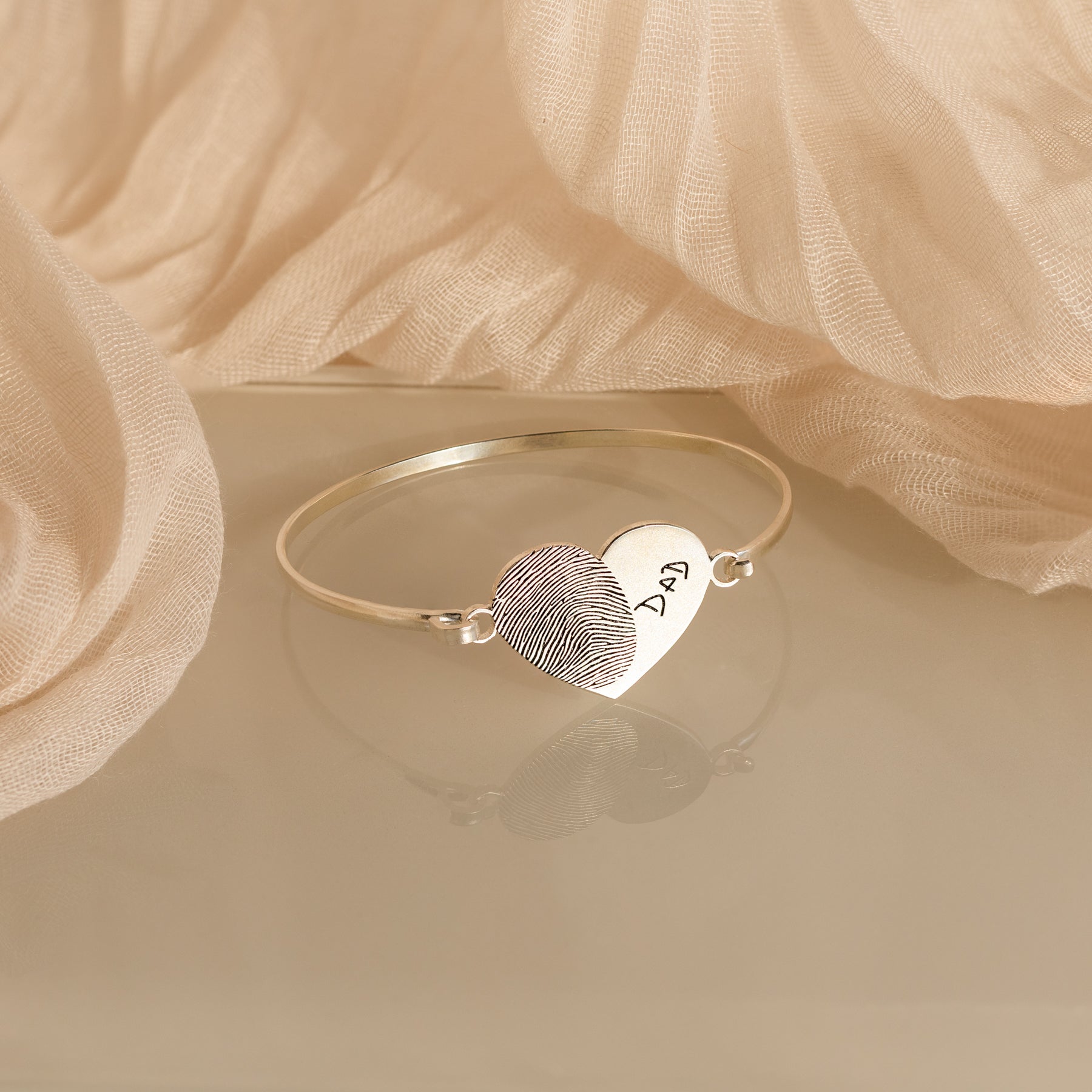 The Fingerprint Heart Bangle is a silver bracelet with a heart charm engraved with a fingerprint and "Dad," making it a unique personalized gift.