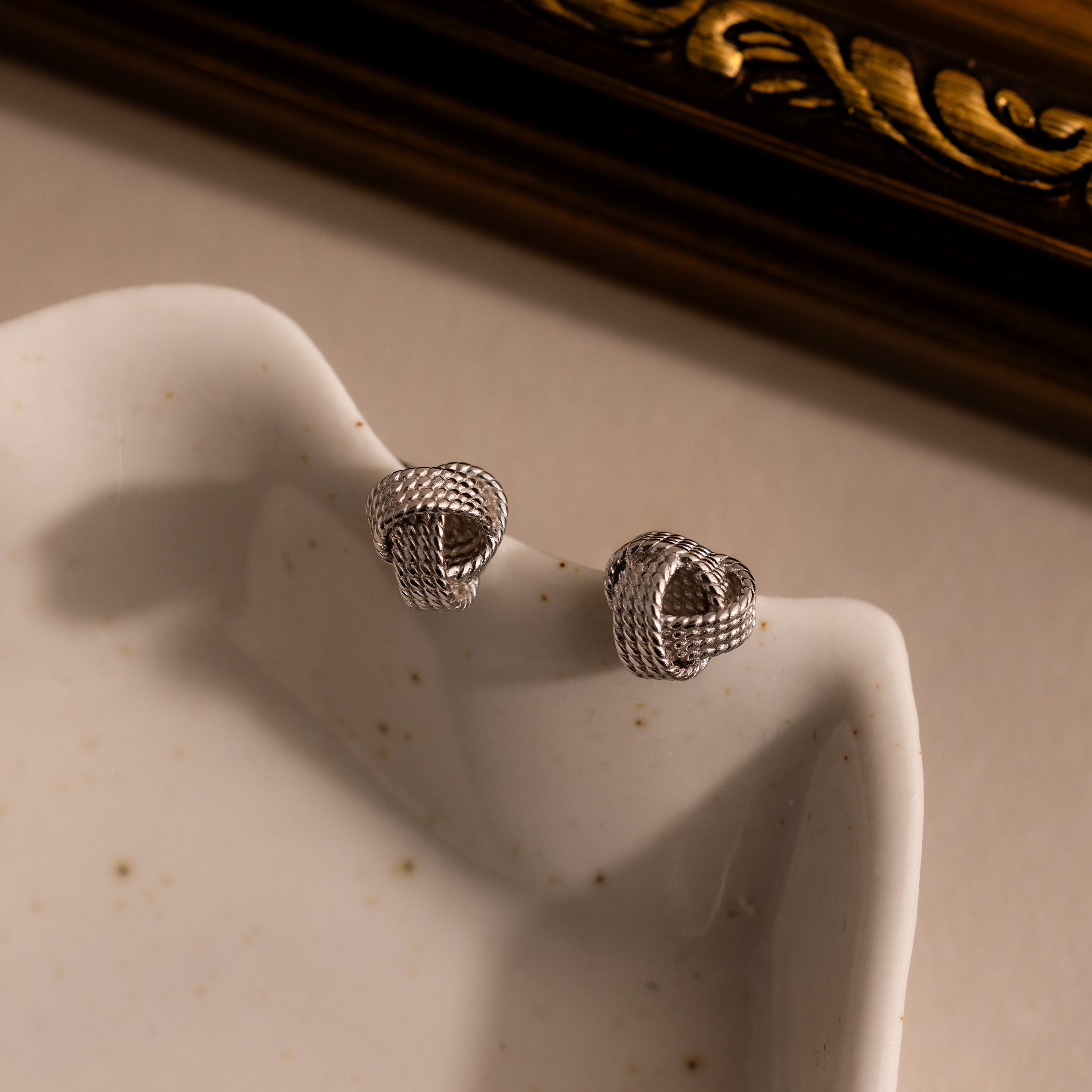 Rope Knot Studs—a pair of silver knot-shaped earrings displayed on a white ceramic dish near a gold frame—perfect for adding timeless elegance to your jewelry collection.
