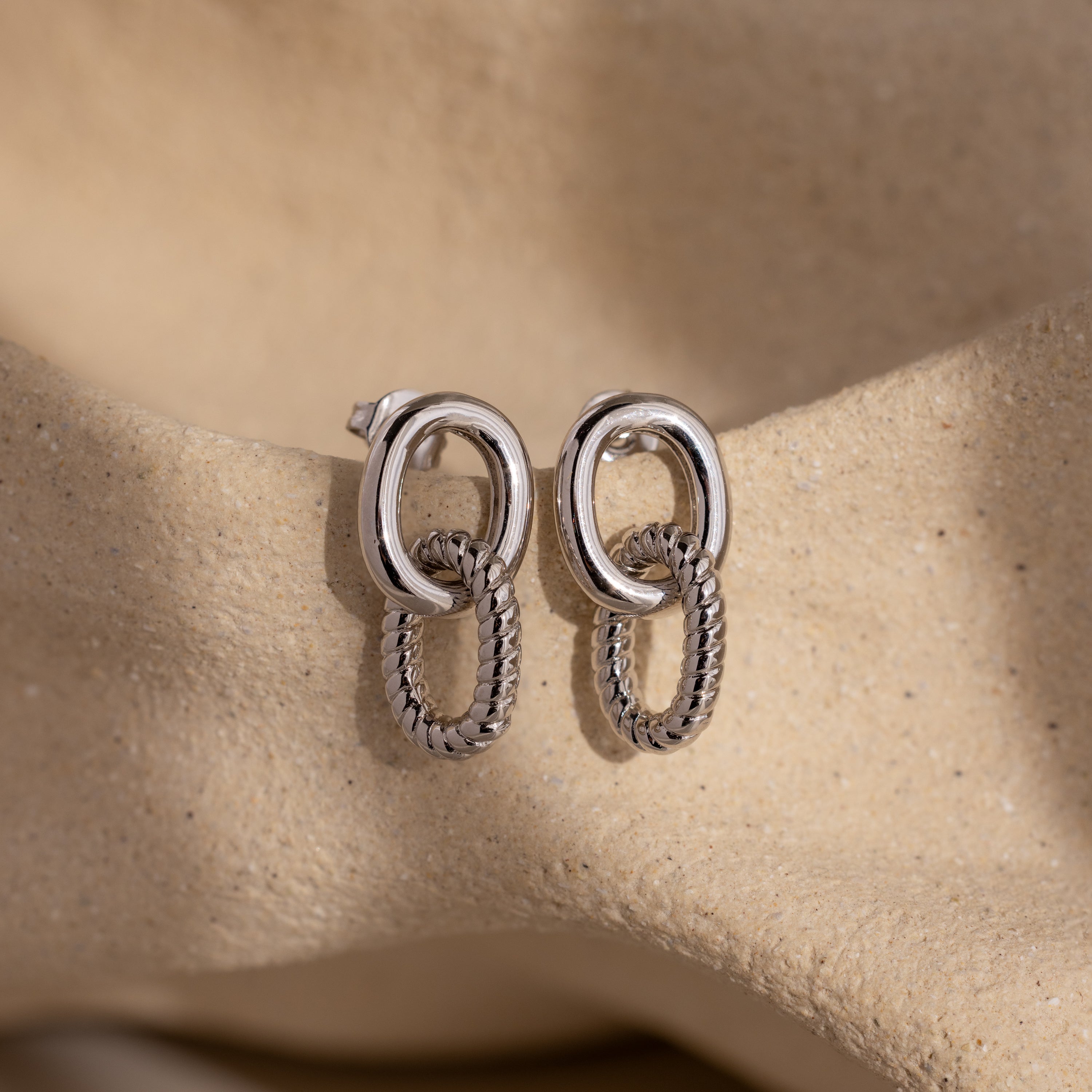 Twisted Link Earrings in White Gold, featuring smooth and textured chains, rest on a soft beige sandy background—modern earrings that embody timeless elegance.