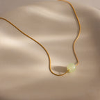 Close-up of a delicate gold chain necklace featuring a single round green jade bead, displayed against a neutral surface.
