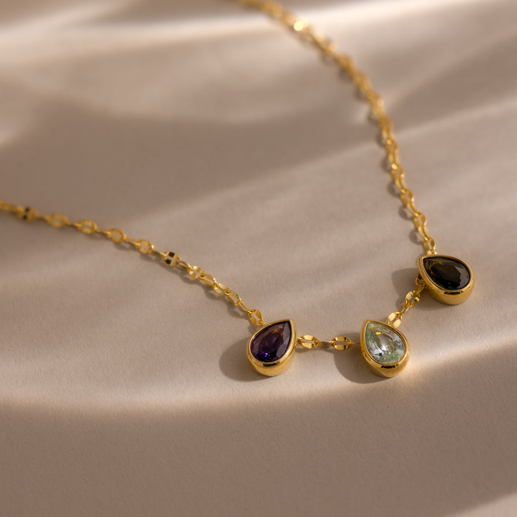 The Multiple Teardrop Birthstone Necklace features three teardrop-shaped gemstones, ideal for personalized jewelry, displayed on a sunlit surface.