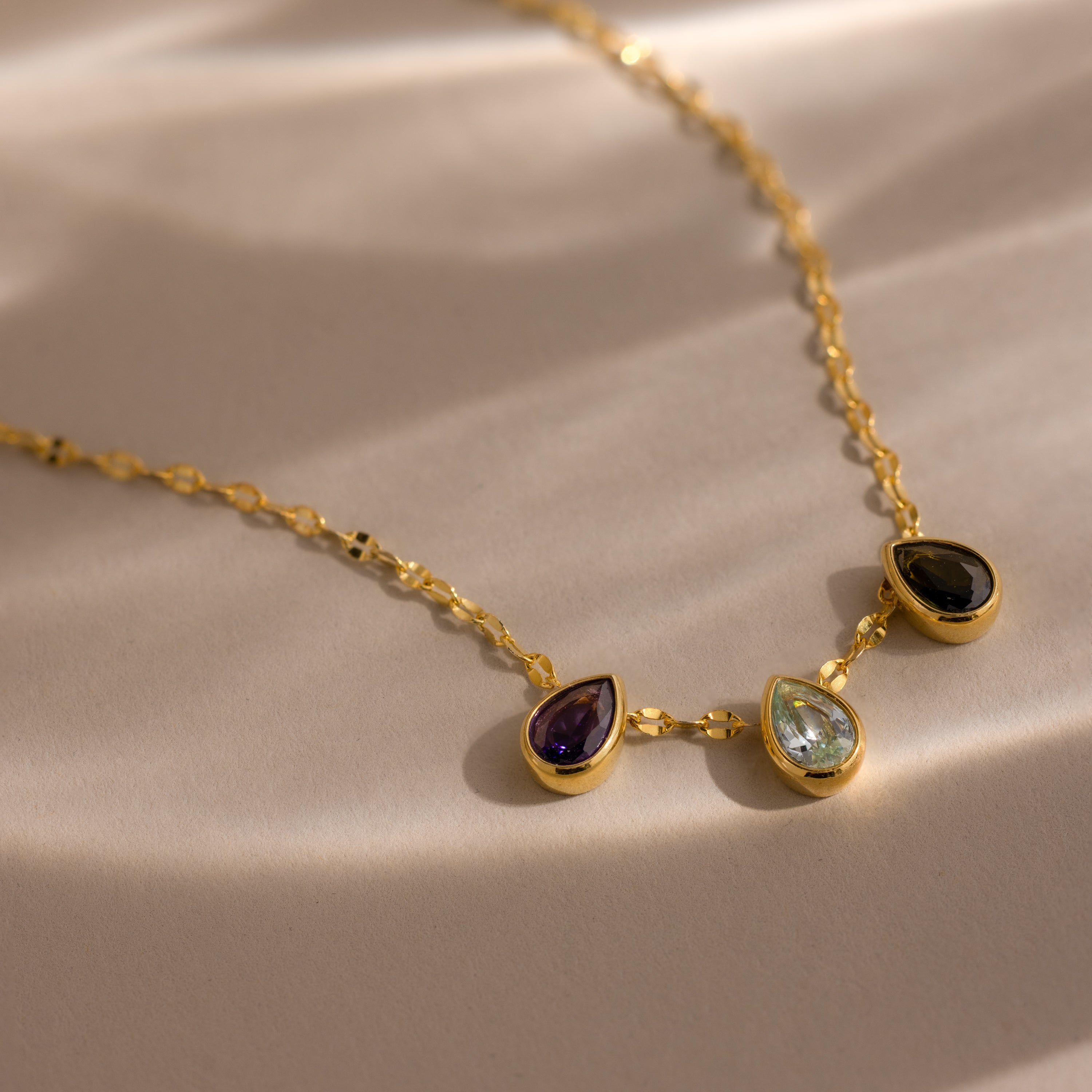 The Multiple Teardrop Birthstone Necklace features three teardrop-shaped gemstones, ideal for personalized jewelry, displayed on a sunlit surface.