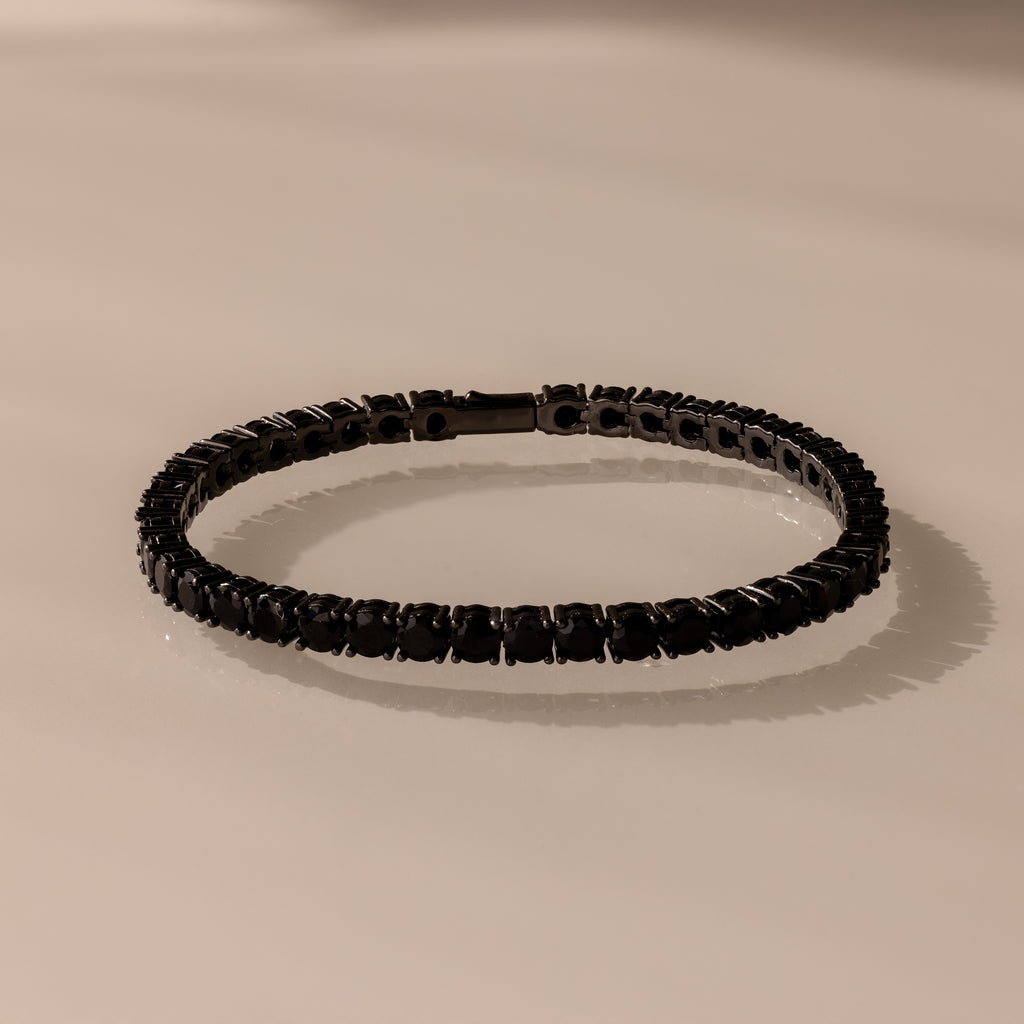 The Onyx Tennis Bracelet features black CZ diamonds and dark gemstones, elegantly displayed on a light beige surface.