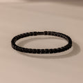 The Onyx Tennis Bracelet features black CZ diamonds and dark gemstones, elegantly displayed on a light beige surface.