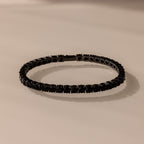 The Onyx Tennis Bracelet features black CZ diamonds and dark gemstones, elegantly displayed on a light beige surface.