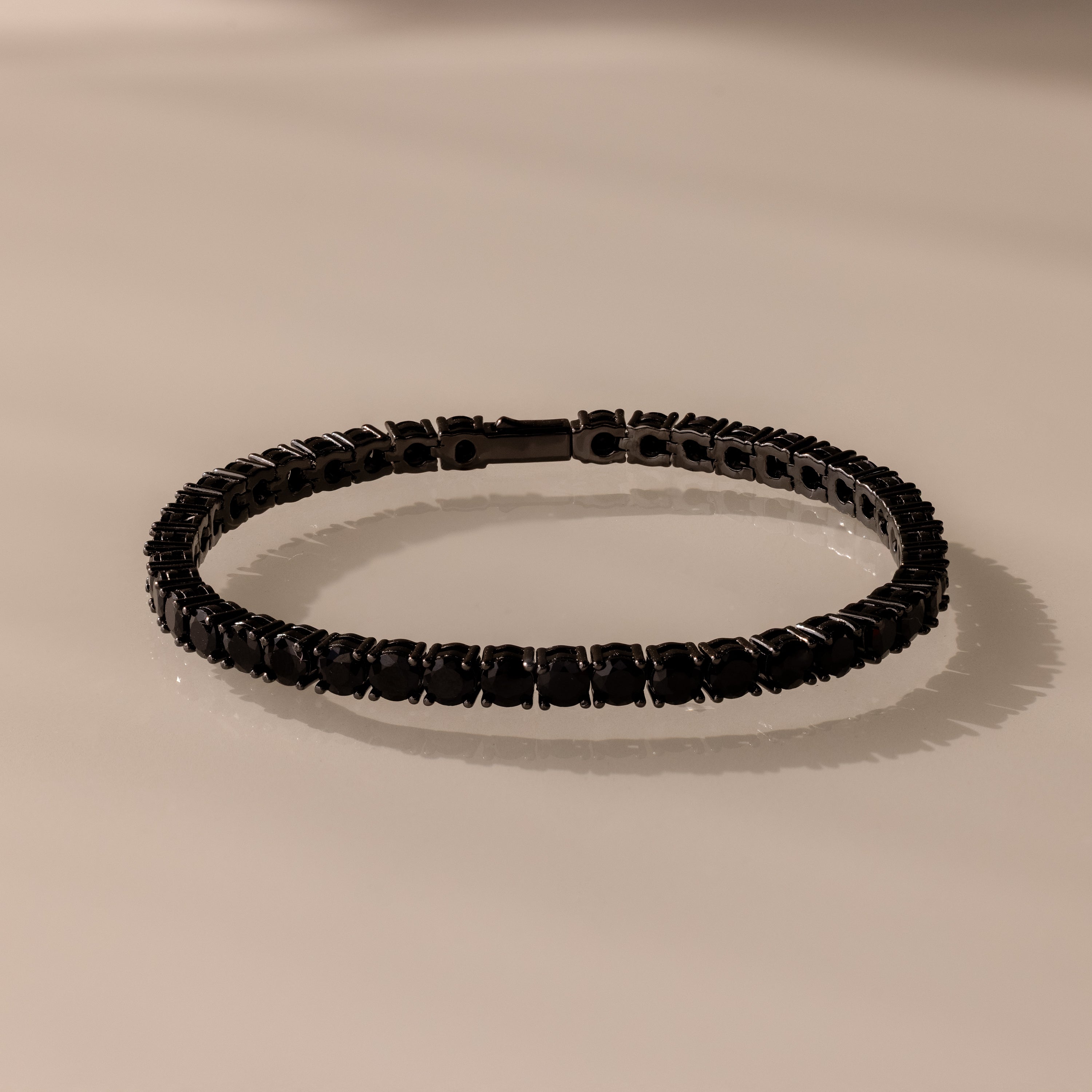 The Onyx Tennis Bracelet features black CZ diamonds and dark gemstones, elegantly displayed on a light beige surface.