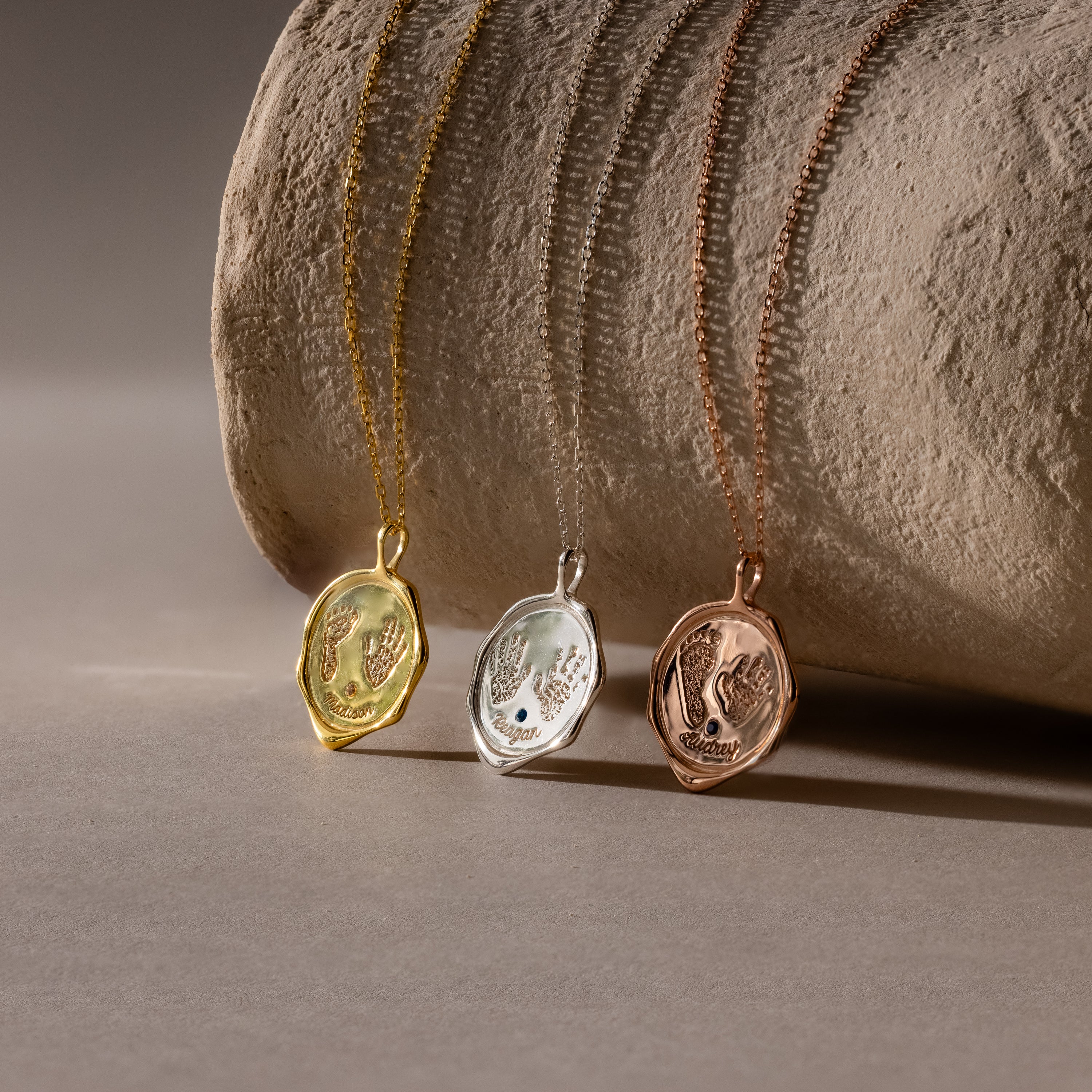 The Baby Hand & Footprint Necklace is shown in gold, silver, and rose gold finishes—ideal for personalized baby jewelry—displayed on a textured cylindrical surface.