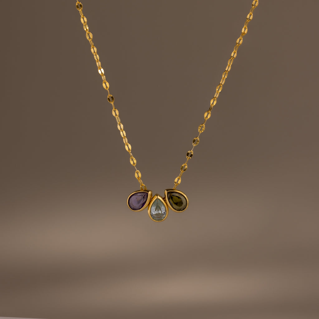 The Multiple Teardrop Birthstone Necklace in 18K Gold features a textured chain with three gemstone pendants—purple, clear teardrop, and green—for a personalized touch to any outfit.