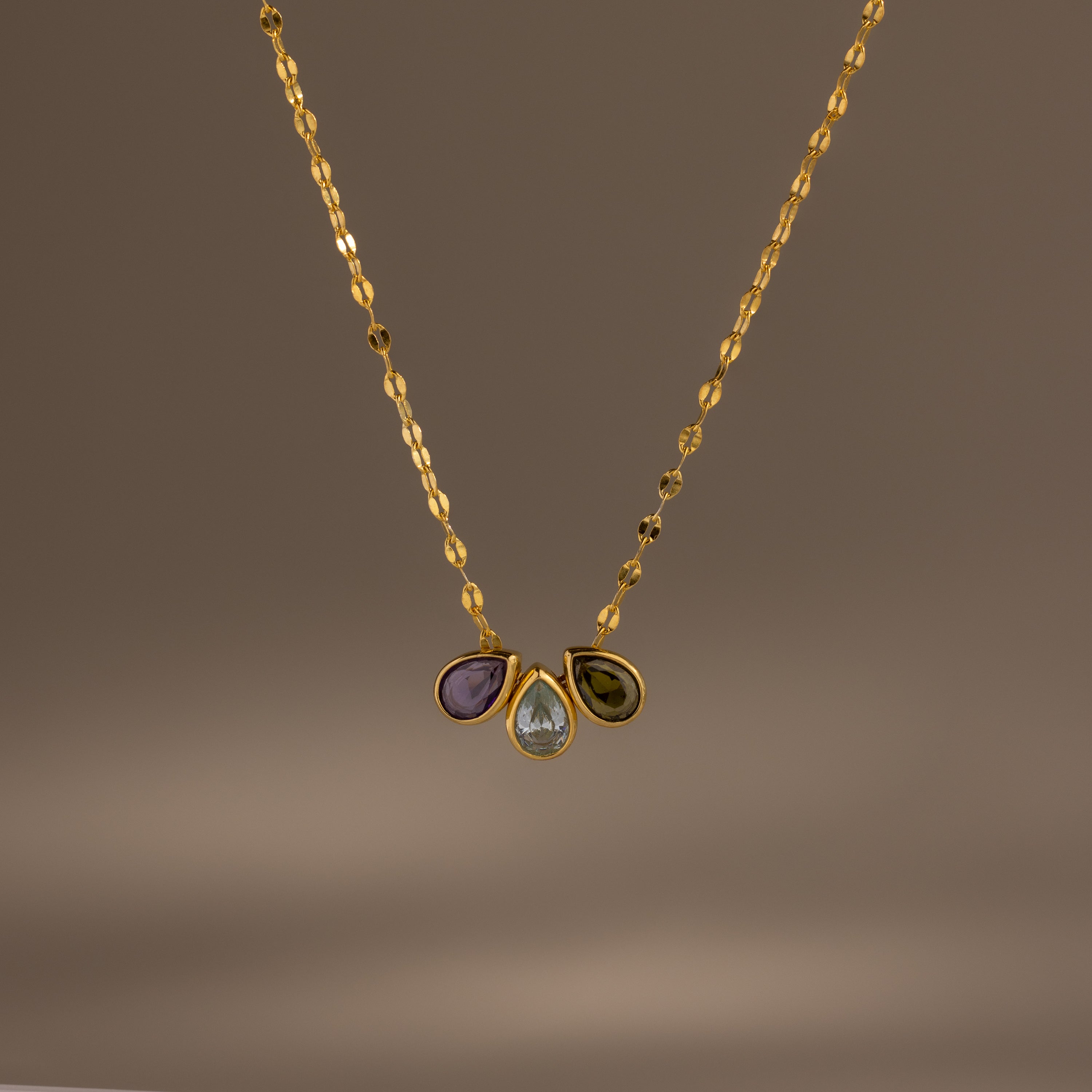 The Multiple Teardrop Birthstone Necklace in 18K Gold features a textured chain with three gemstone pendants—purple, clear teardrop, and green—for a personalized touch to any outfit.