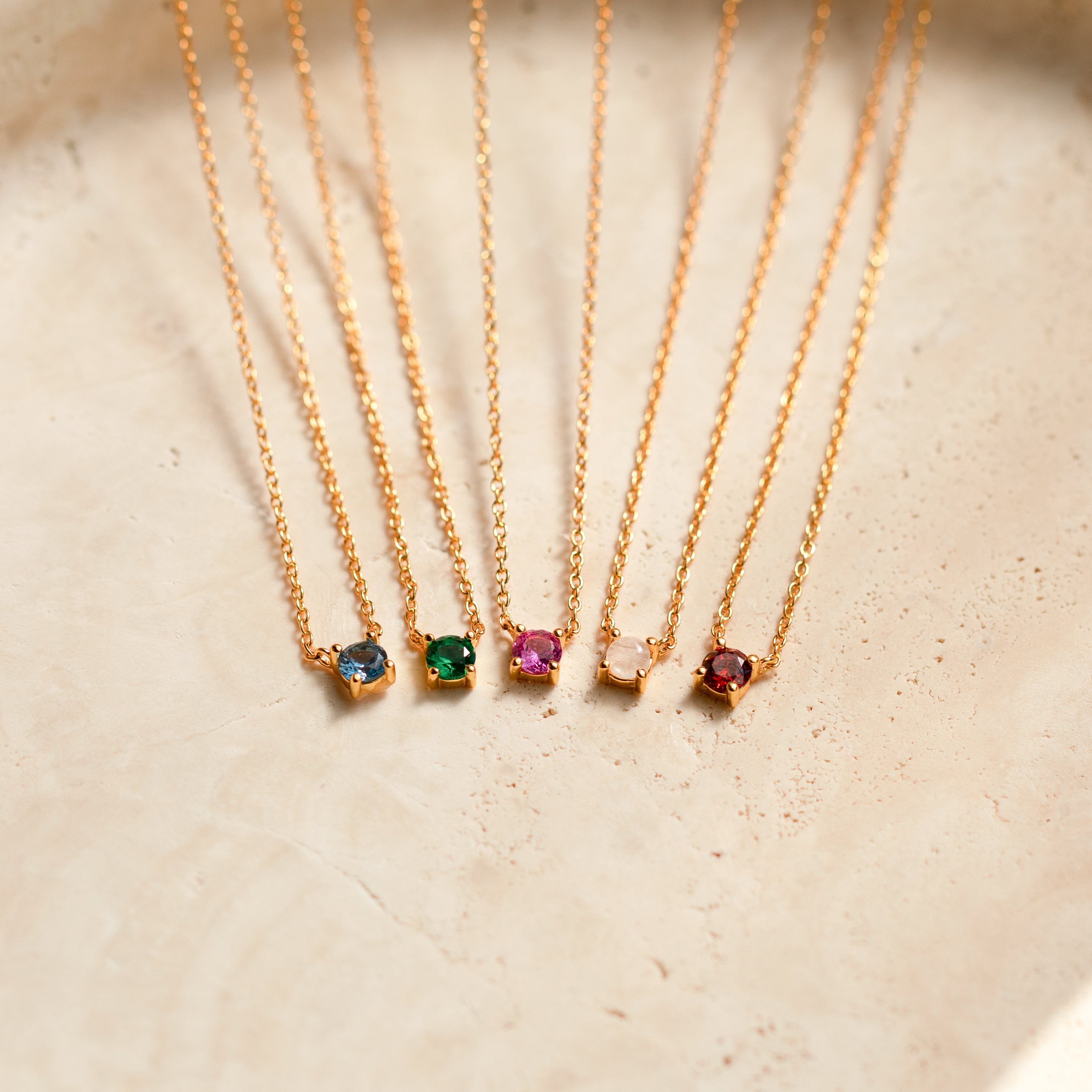 Five dainty necklaces with gold chains and circle birthstone gems laid on a cream ceramic background. Starting left to right the gemstones featured show zircon, emerald, ruby, moonstone, and garnet.