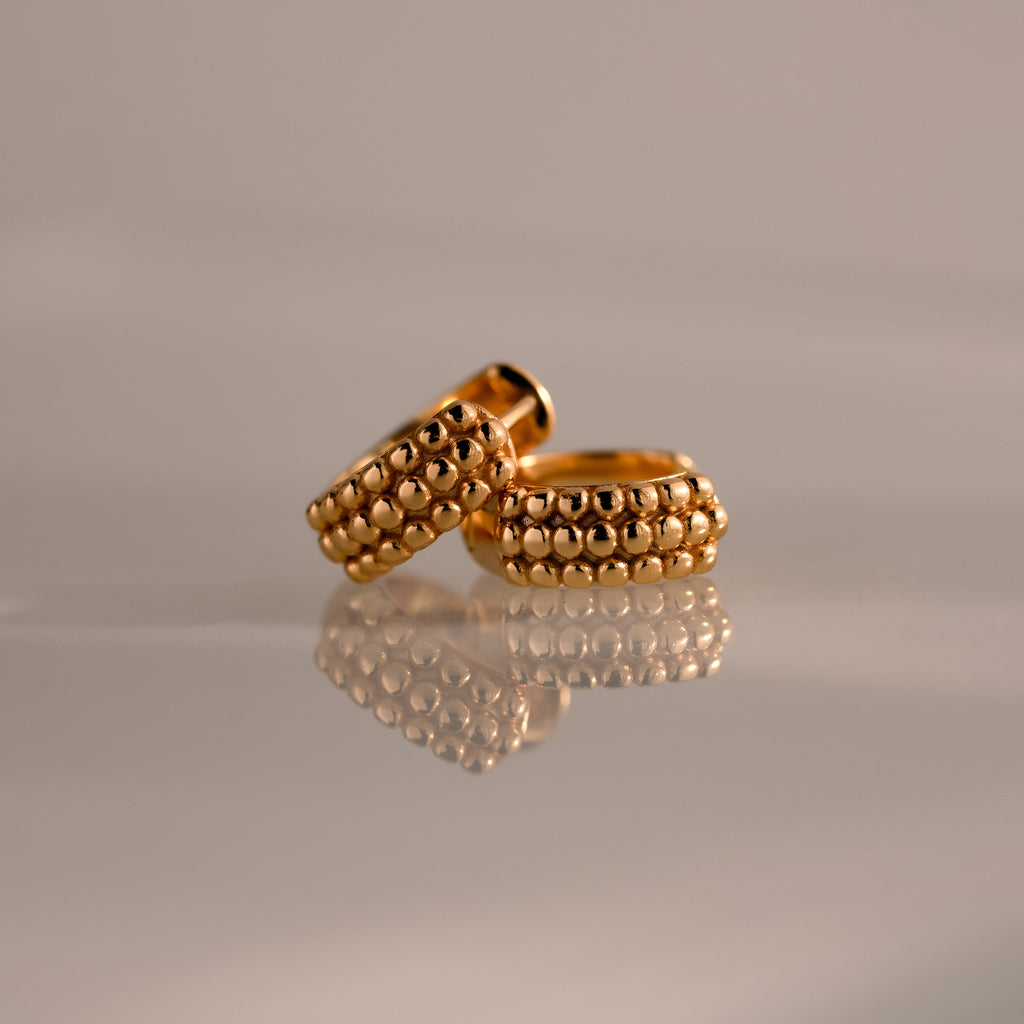 The Beaded Chunky Hoops in 18K Gold, featuring beaded detailing, are displayed on a reflective surface against a soft, neutral background.