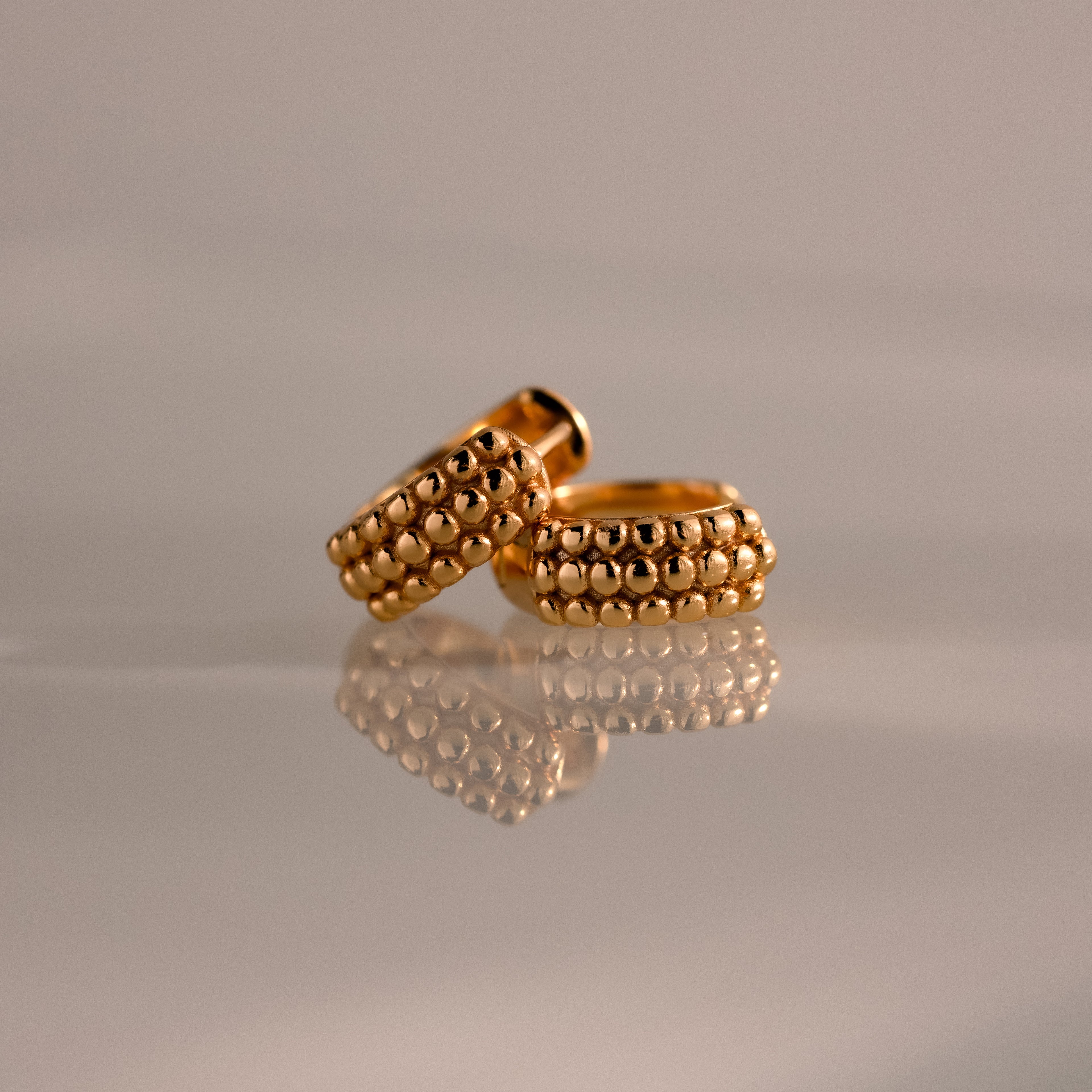 The Beaded Chunky Hoops in 18K Gold, featuring beaded detailing, are displayed on a reflective surface against a soft, neutral background.