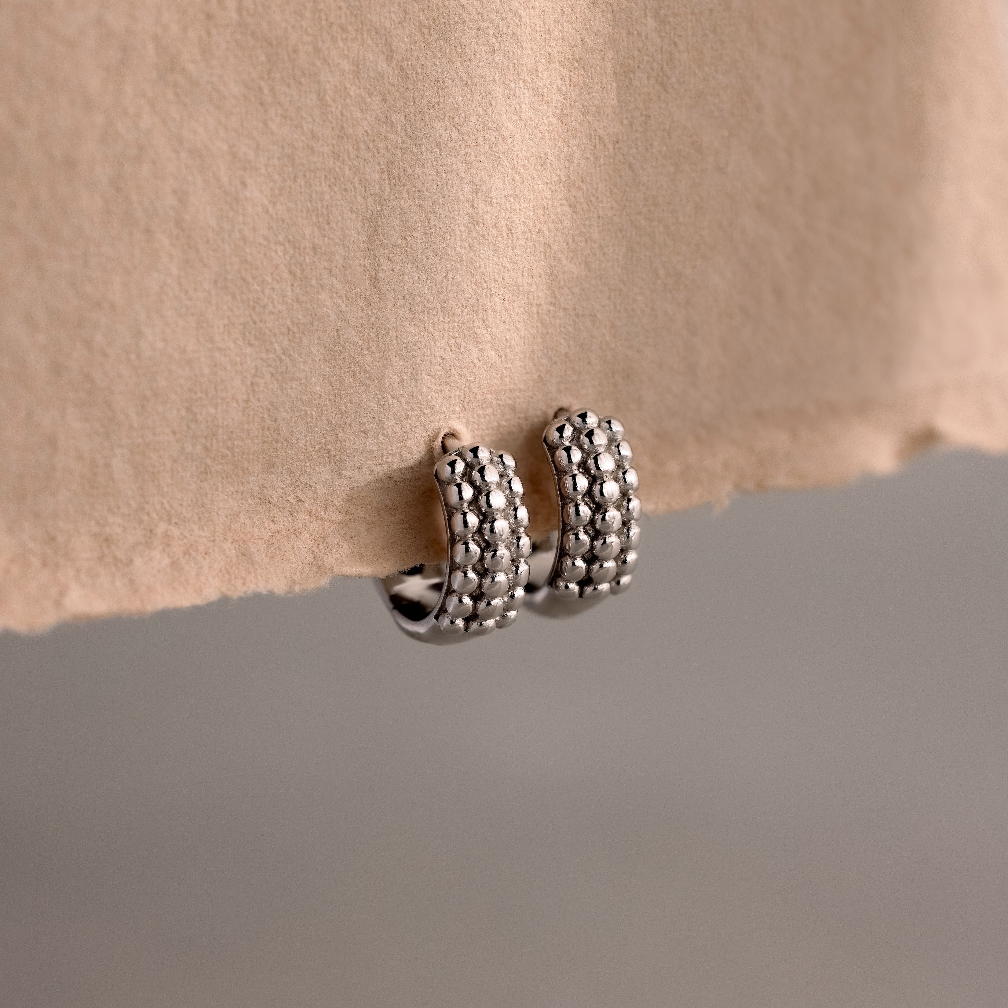 Beaded Chunky Hoops in Sterling Silver rest on a beige paper edge, showcasing a modern, textured design ideal for elevating any contemporary style.