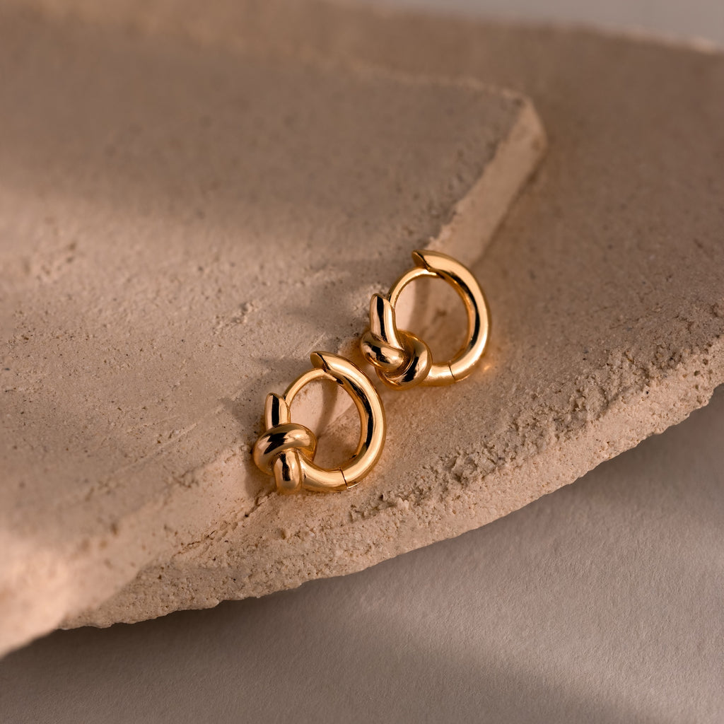 Two Love Knot Huggies with intricate knot details rest on a textured beige stone surface, representing lasting bonds and making a thoughtful gift.