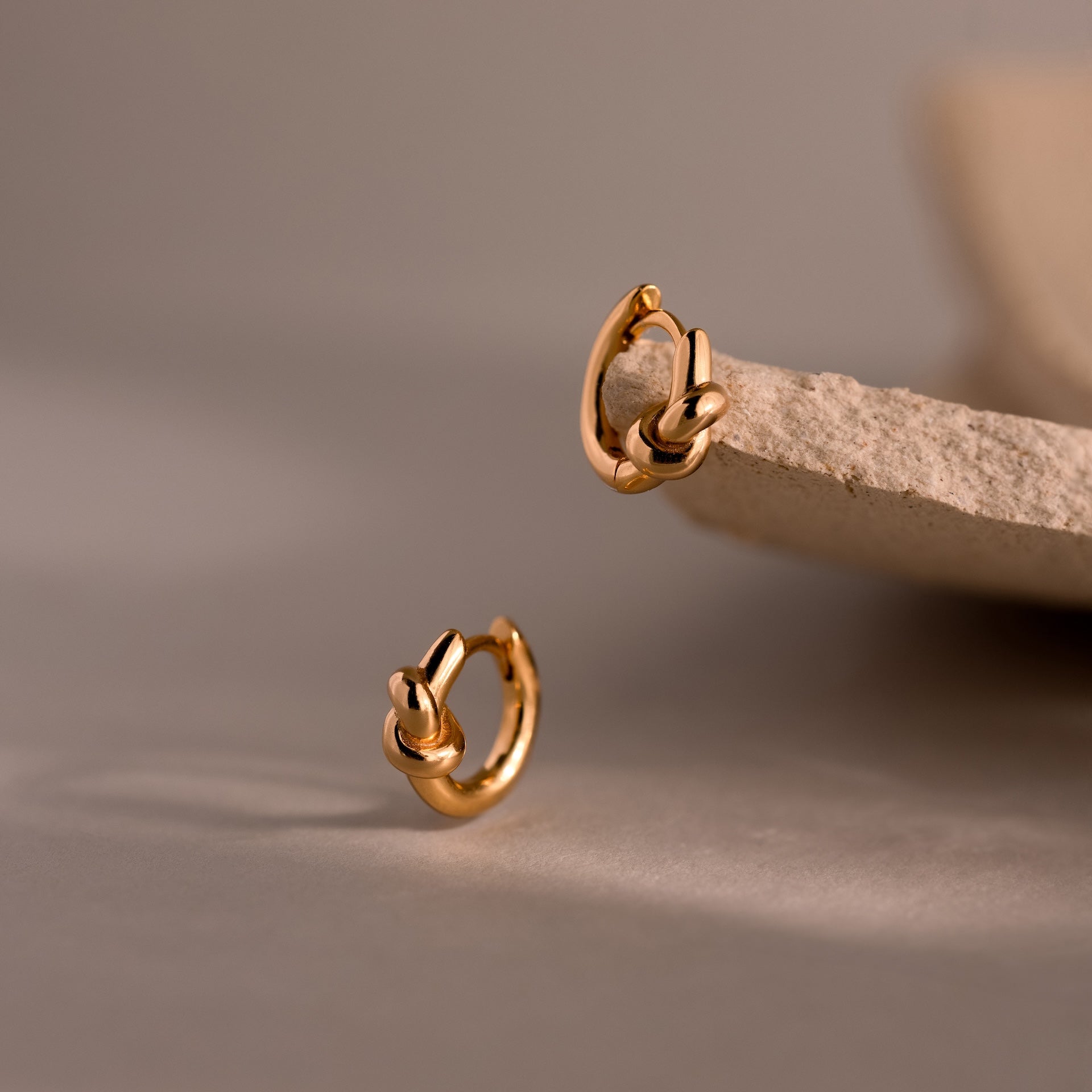 Love Knot Huggies in 18K Gold—two gold hoop earrings with knot details, one on a stone and one on a neutral surface—symbolize lasting bonds and make a meaningful gift for someone special.