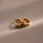 Two Love Knot Huggies in 18K Gold with elegant knot designs rest on a soft beige surface under gentle lighting—an ideal meaningful gift to celebrate lasting bonds.