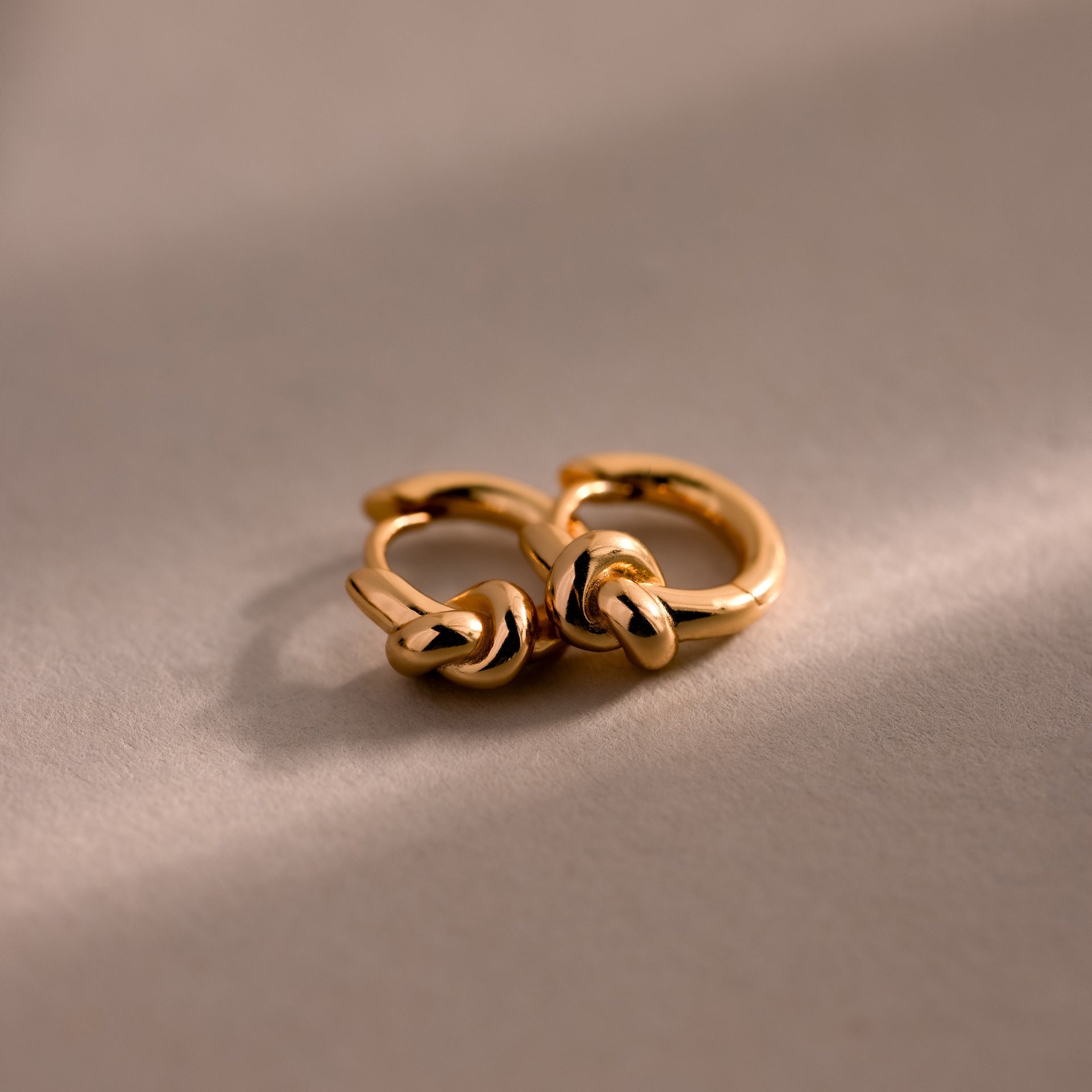 Two Love Knot Huggies in 18K Gold with elegant knot designs rest on a soft beige surface under gentle lighting—an ideal meaningful gift to celebrate lasting bonds.