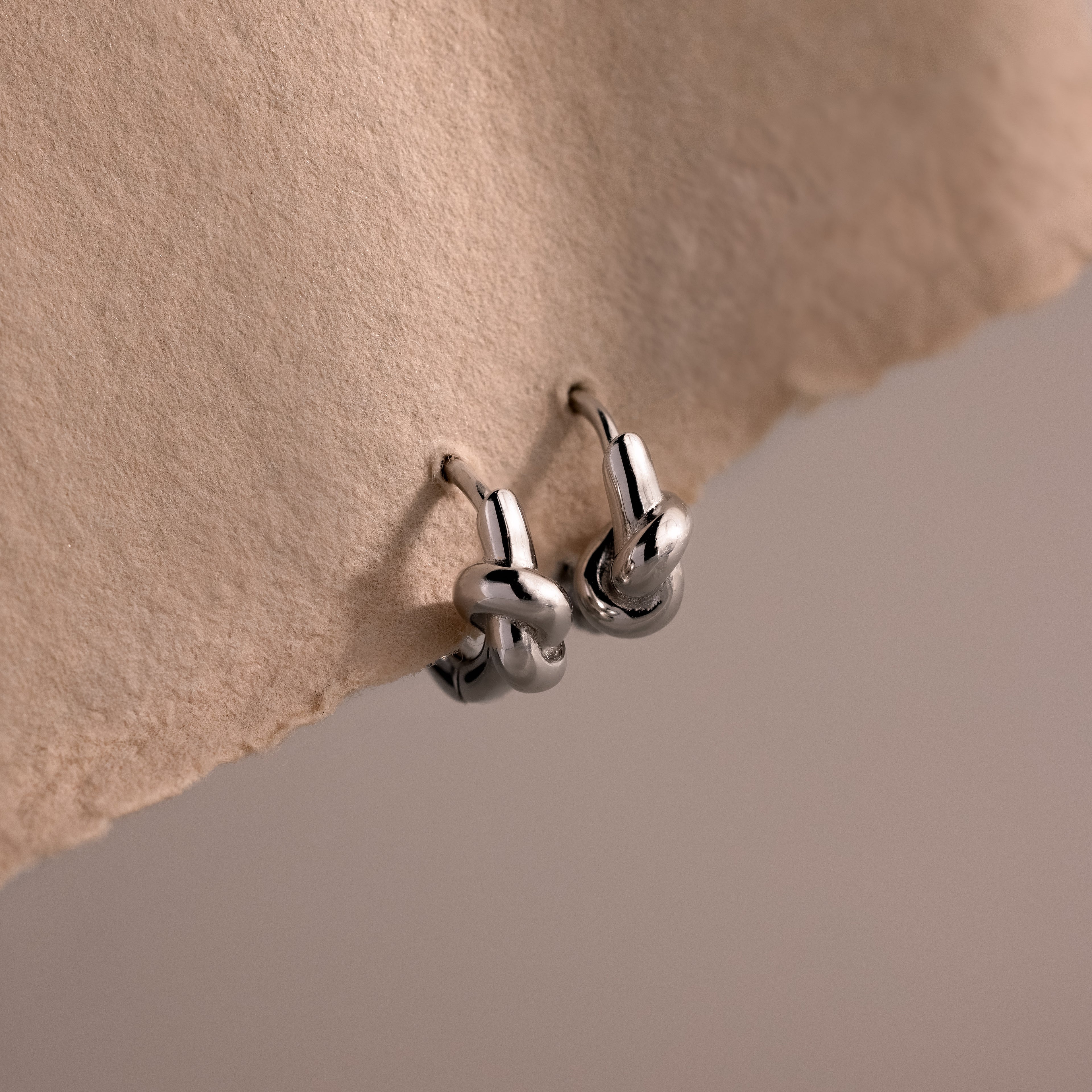 Love Knot Huggies in Sterling Silver are shown on textured beige paper against a soft, neutral background—an ideal, meaningful gift to celebrate lasting bonds.