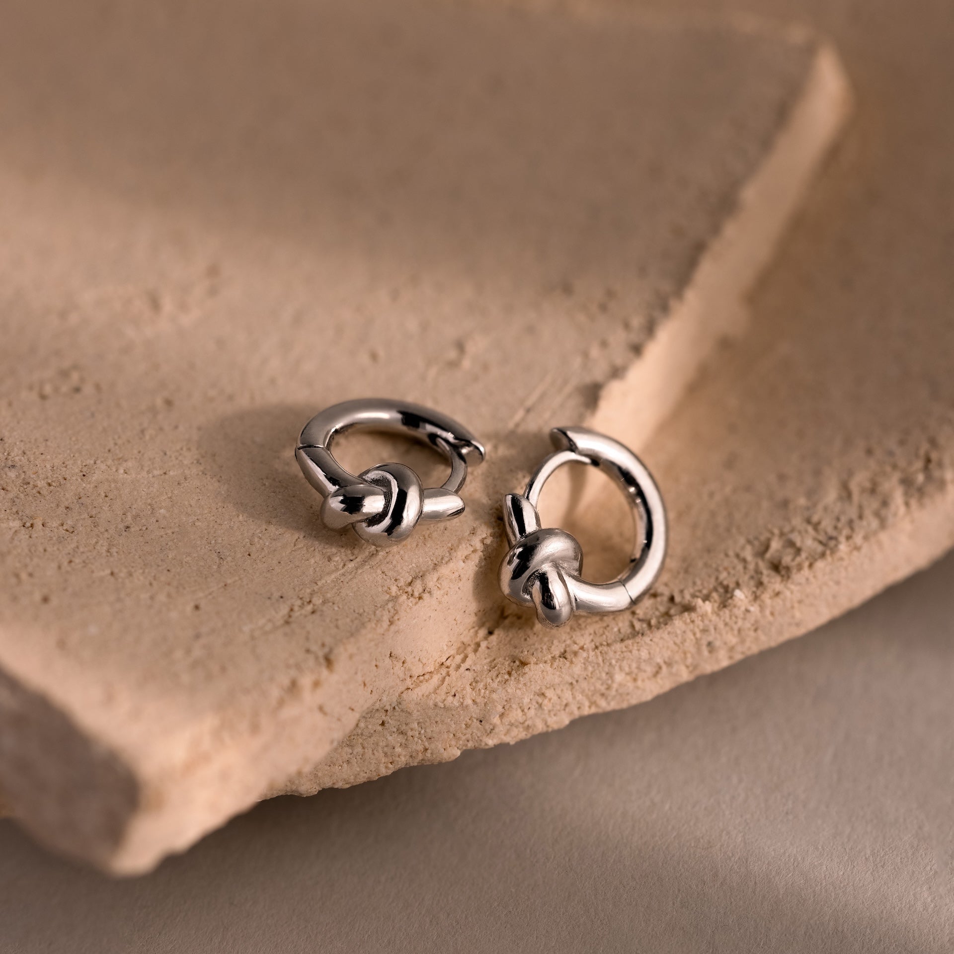Two Love Knot Huggies in sterling silver rest on textured beige stone slabs, symbolizing lasting bonds—an elegant and meaningful gift.