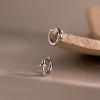 Two Love Knot Huggies in Sterling Silver are displayed on a beige stone surface with a soft neutral background, symbolizing lasting bonds and making a meaningful gift.