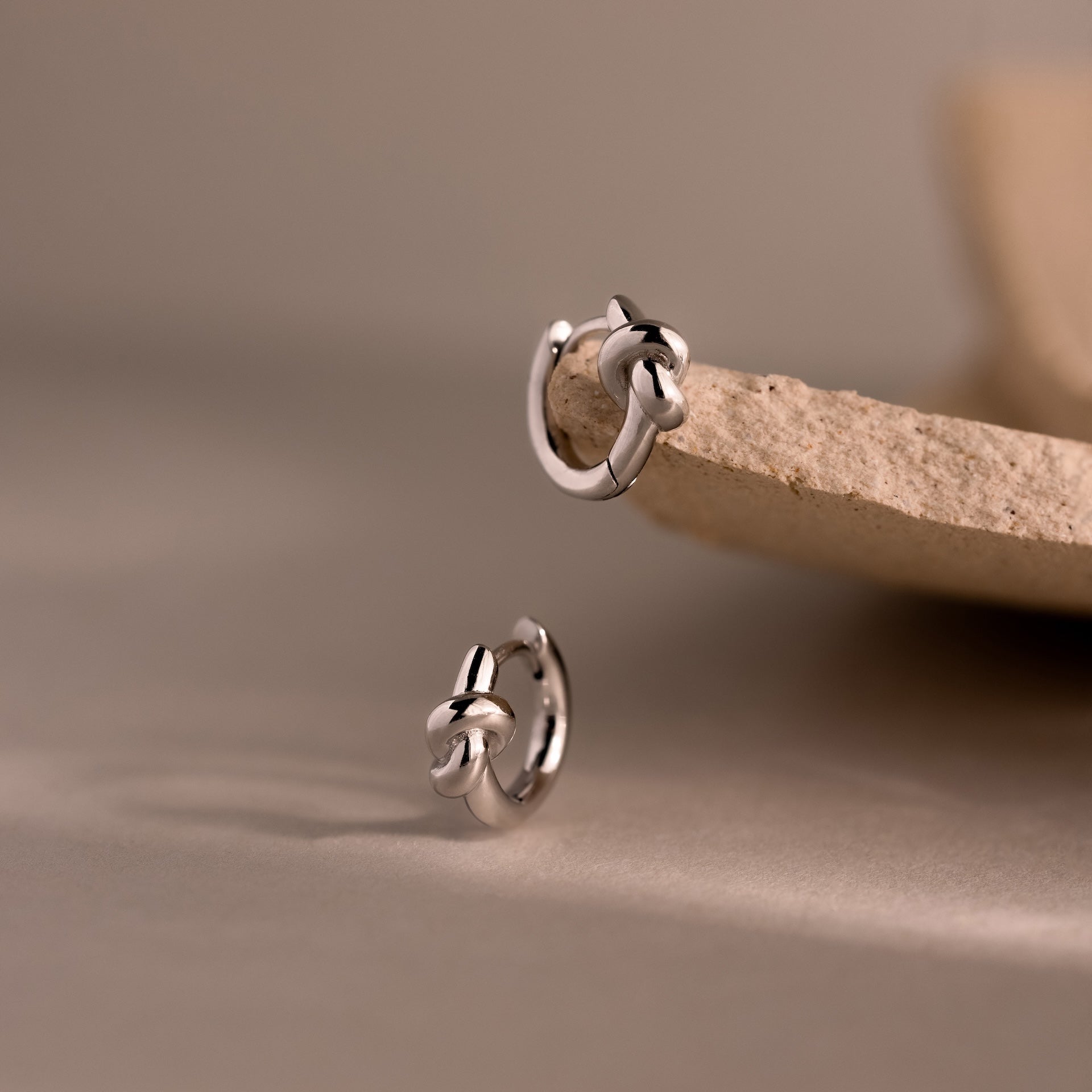 Two Love Knot Huggies in Sterling Silver are displayed on a beige stone surface with a soft neutral background, symbolizing lasting bonds and making a meaningful gift.