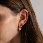 Close-up of a woman’s ear with three gold earrings, including Love Knot Huggies in 18K Gold—an elegant gift symbolizing lasting bonds, her brown hair tucked behind her ear.