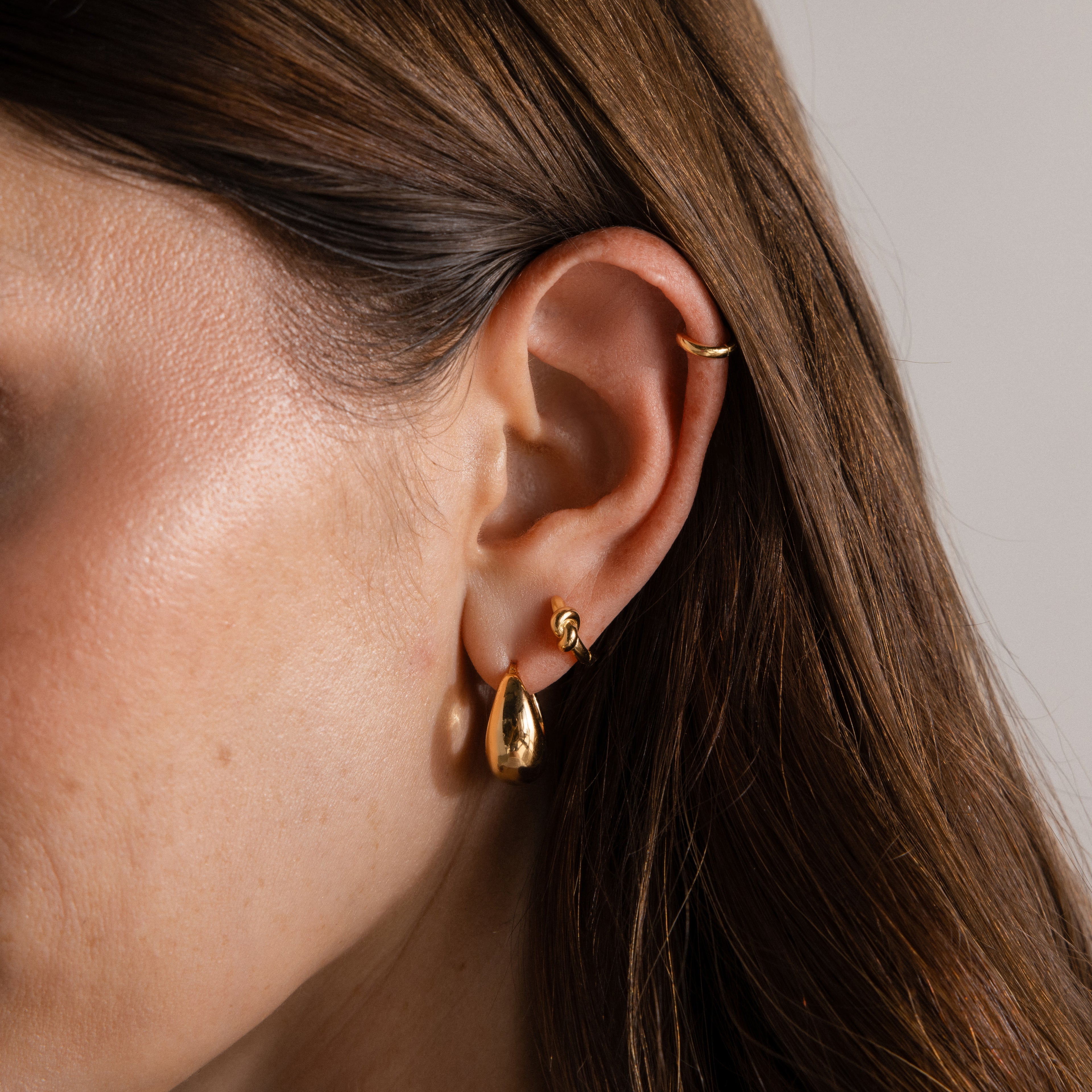 Close-up of a woman’s ear with three gold earrings, including Love Knot Huggies in 18K Gold—an elegant gift symbolizing lasting bonds, her brown hair tucked behind her ear.