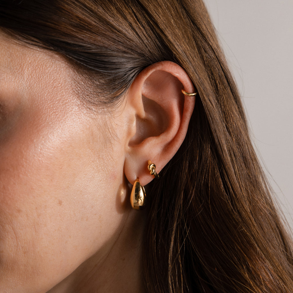 Close-up of a woman's ear wearing three gold earrings: a hoop, a small stud, and the Love Knot Huggies—teardrop-shaped earrings perfect for celebrating lasting bonds or giving as a meaningful gift.