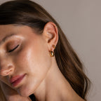 A woman with closed eyes, gently touching her face and wearing three gold Love Knot Huggies earrings, showcases smooth brown hair—a meaningful gift that represents lasting bonds.