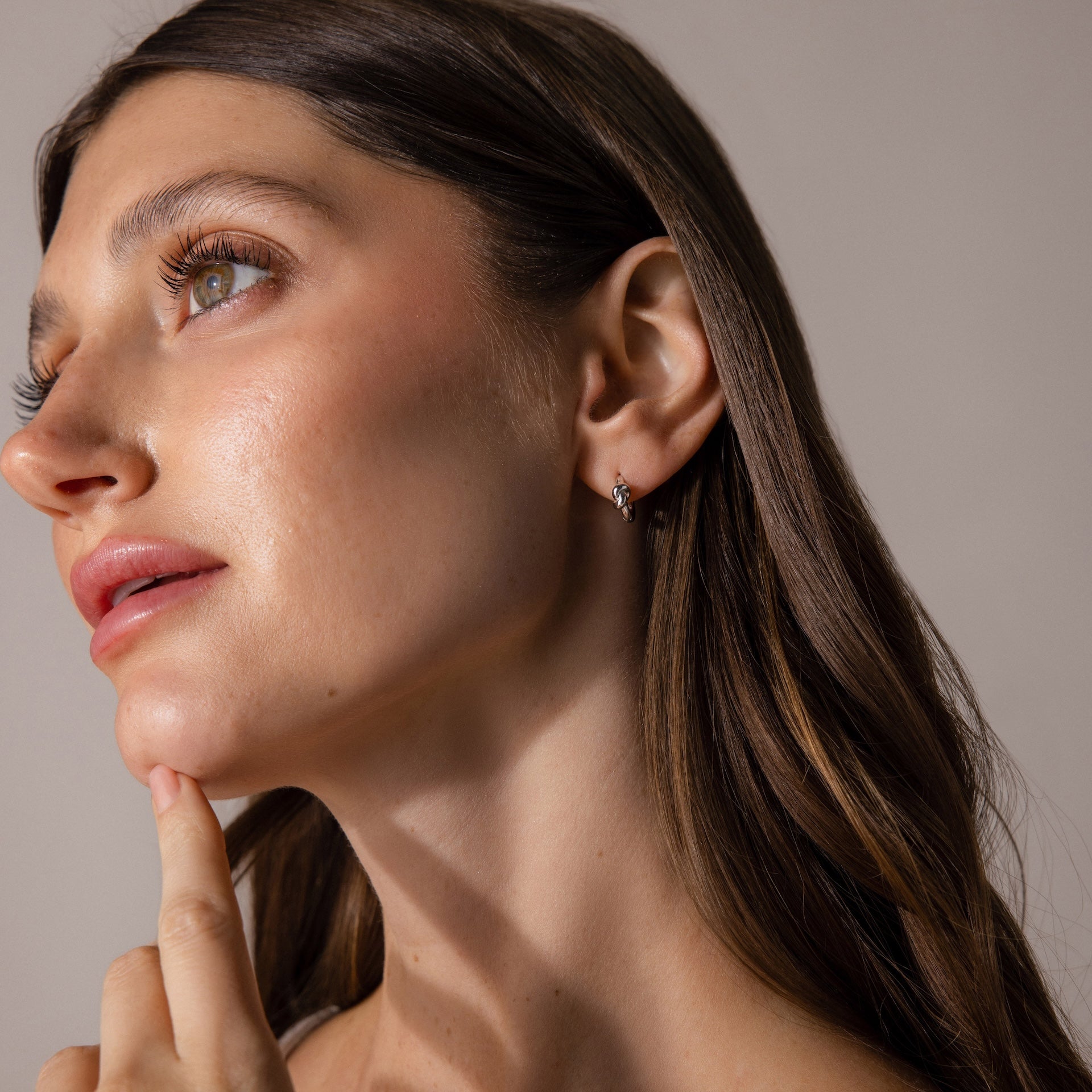 Woman with long brown hair touches her chin and looks up, wearing Love Knot Huggies in Sterling Silver—these earrings symbolize lasting bonds, making them a meaningful gift. Neutral background.
