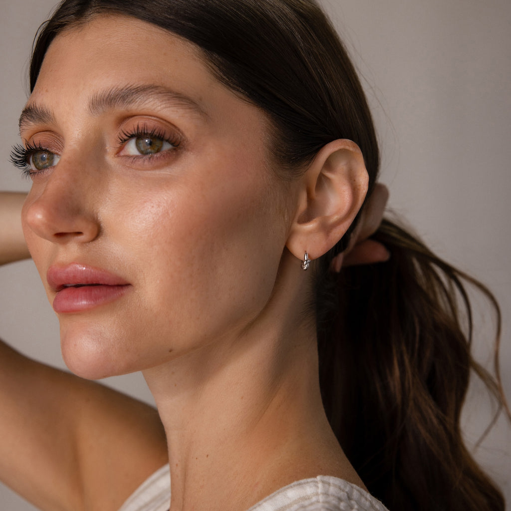 A woman with long brown hair in a ponytail wears Love Knot Huggies, looking off to the side—the elegant earrings symbolize lasting bonds and make a meaningful gift.