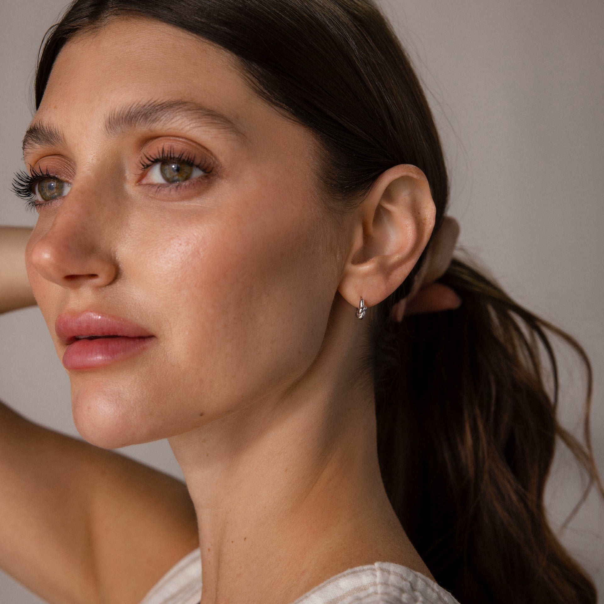 A woman with long brown hair in a ponytail wears Love Knot Huggies, looking off to the side—the elegant earrings symbolize lasting bonds and make a meaningful gift.