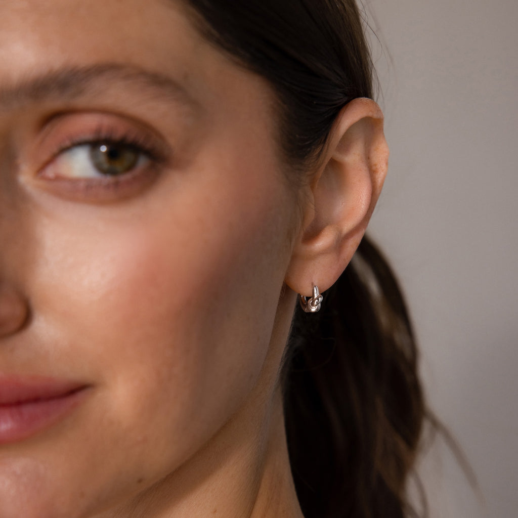 Close-up of a woman with brown hair wearing Love Knot Huggies in Sterling Silver, softly smiling—a meaningful gift that celebrates lasting bonds.