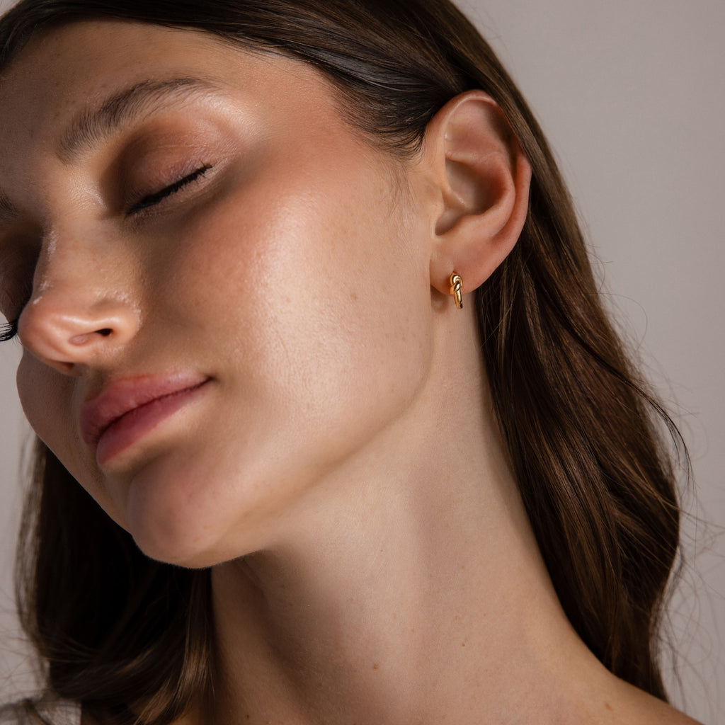 With eyes closed and head tilted, a woman wears the Love Knot Huggies in 18K Gold—an elegant earring and subtle symbol of lasting bonds, making a meaningful gift.