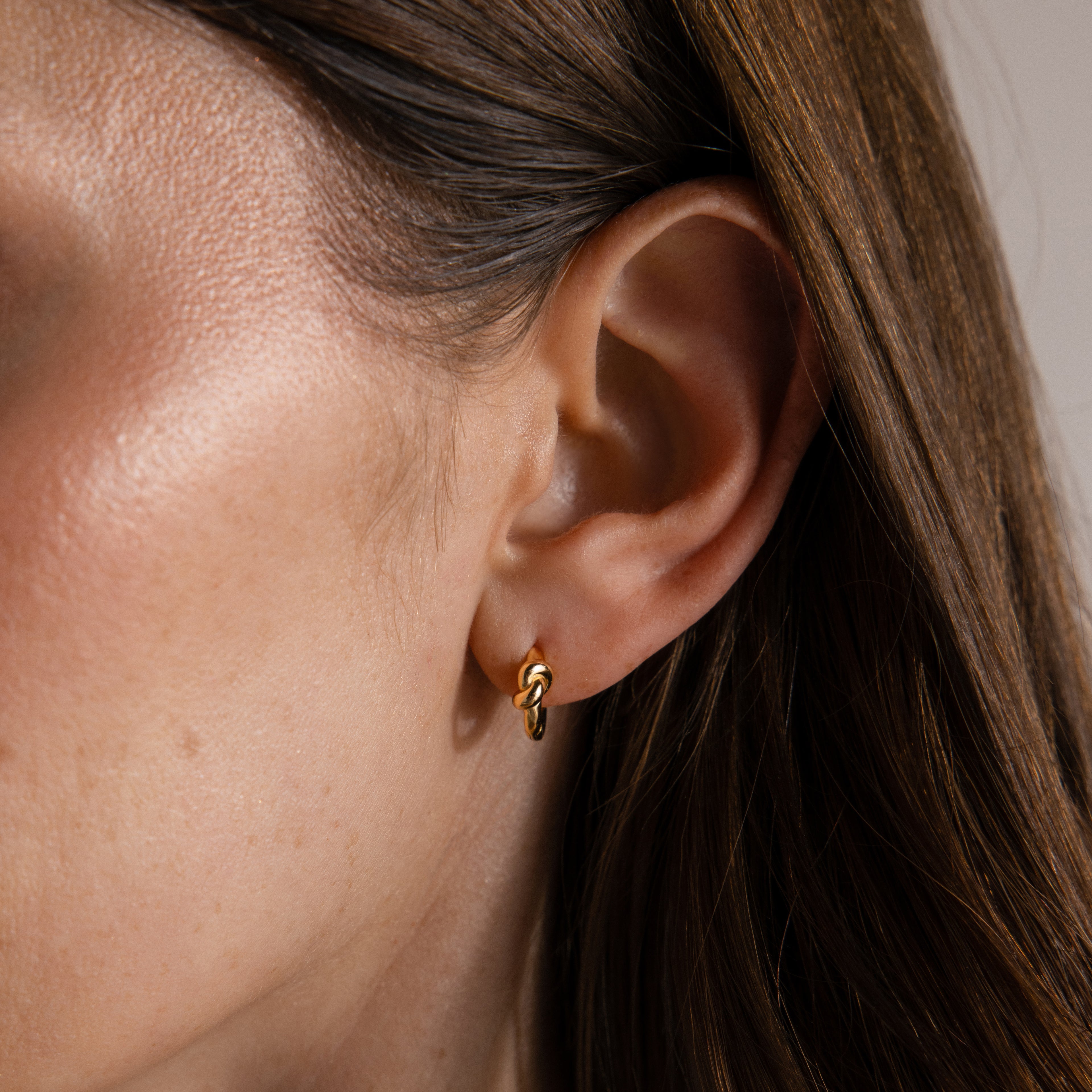 Close-up of a woman's ear with straight brown hair, wearing Love Knot Huggies—small gold earrings symbolizing timeless bonds.