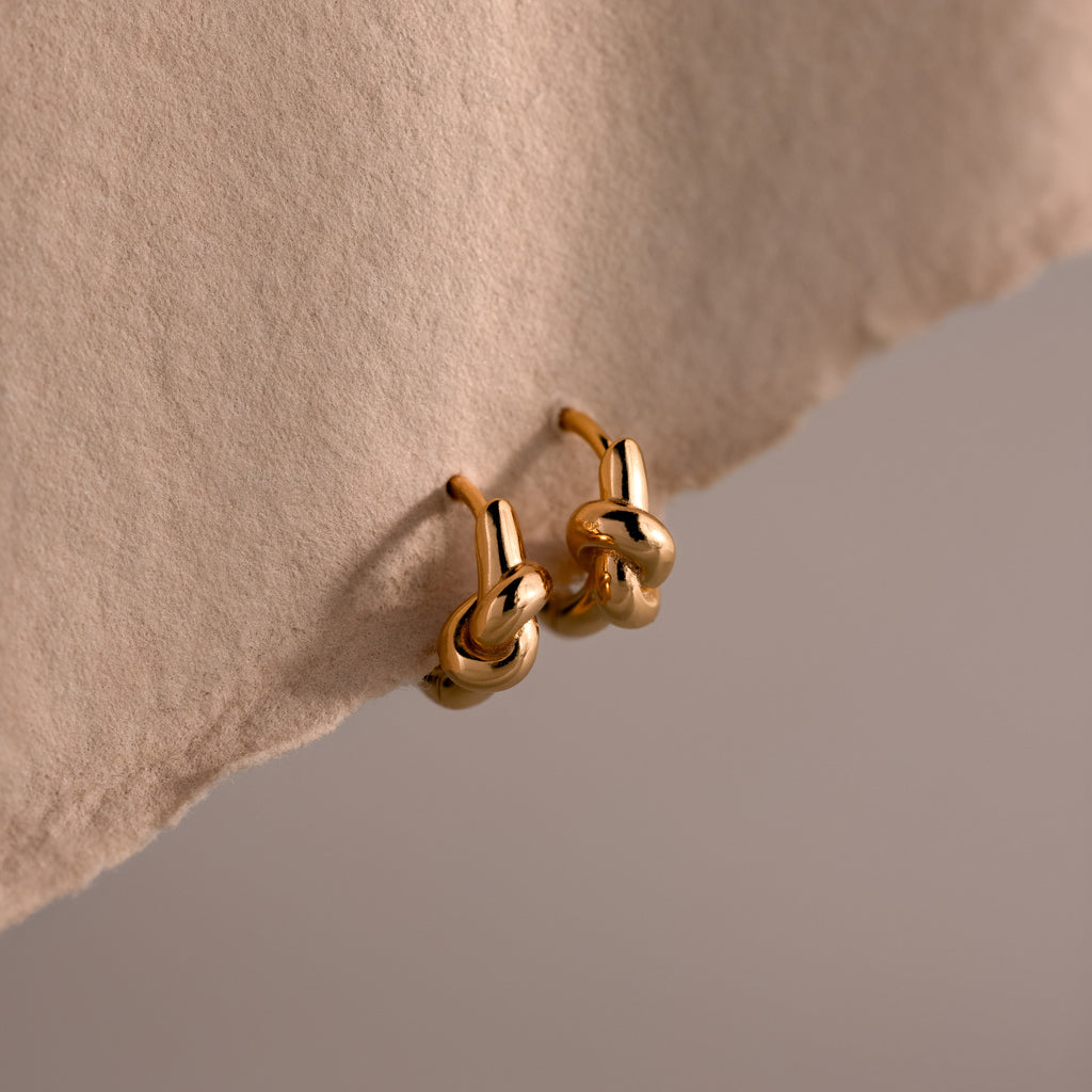 Love Knot Huggies in 18K Gold displayed on textured beige fabric, symbolizing lasting bonds—an elegant and meaningful gift.