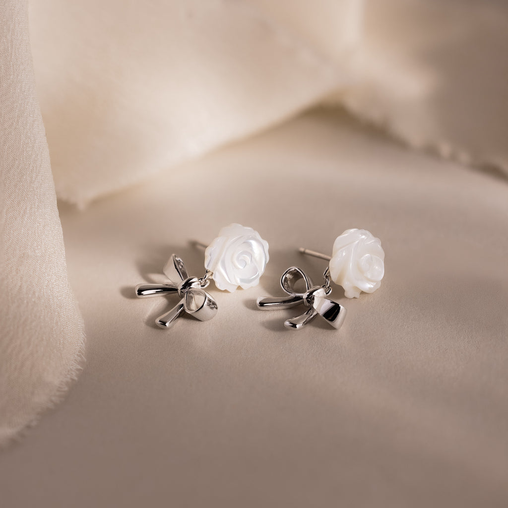White rose and silver bow earrings displayed on a neutral surface.
