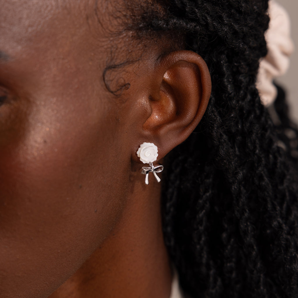Up-close of model wearing white pearl rose stud earring with a dangling silver ribbon bow charm.