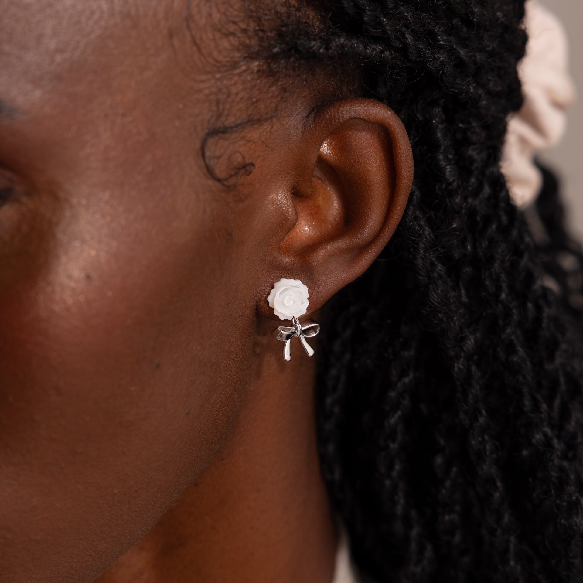 Up-close of model wearing white pearl rose stud earring with a dangling silver ribbon bow charm.