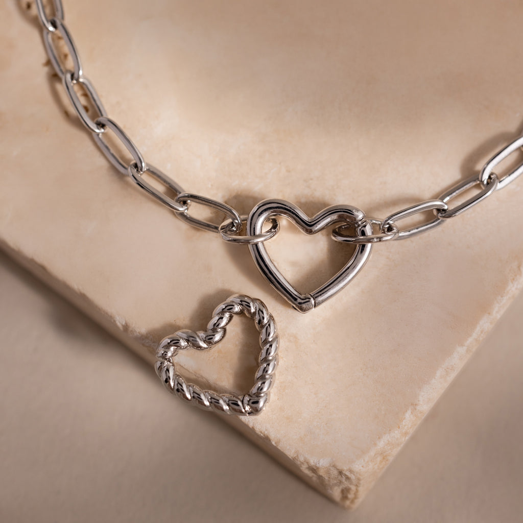 Close-up of a silver heart carabiner bracelet with paperclip chain links, shown with a second twisted heart charm on a stone surface.