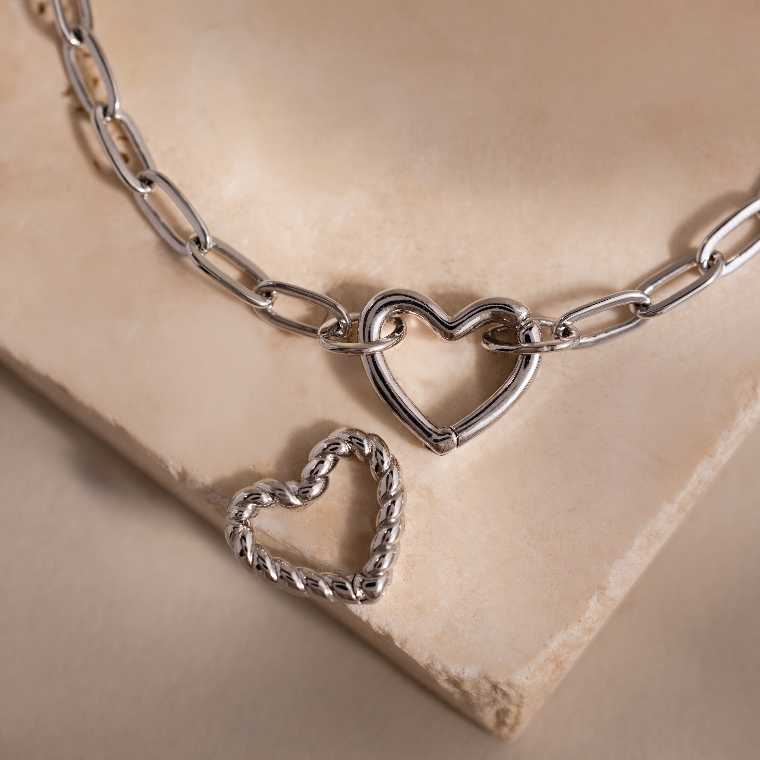 Close-up of a silver heart carabiner bracelet with paperclip chain links, shown with a second twisted heart charm on a stone surface.