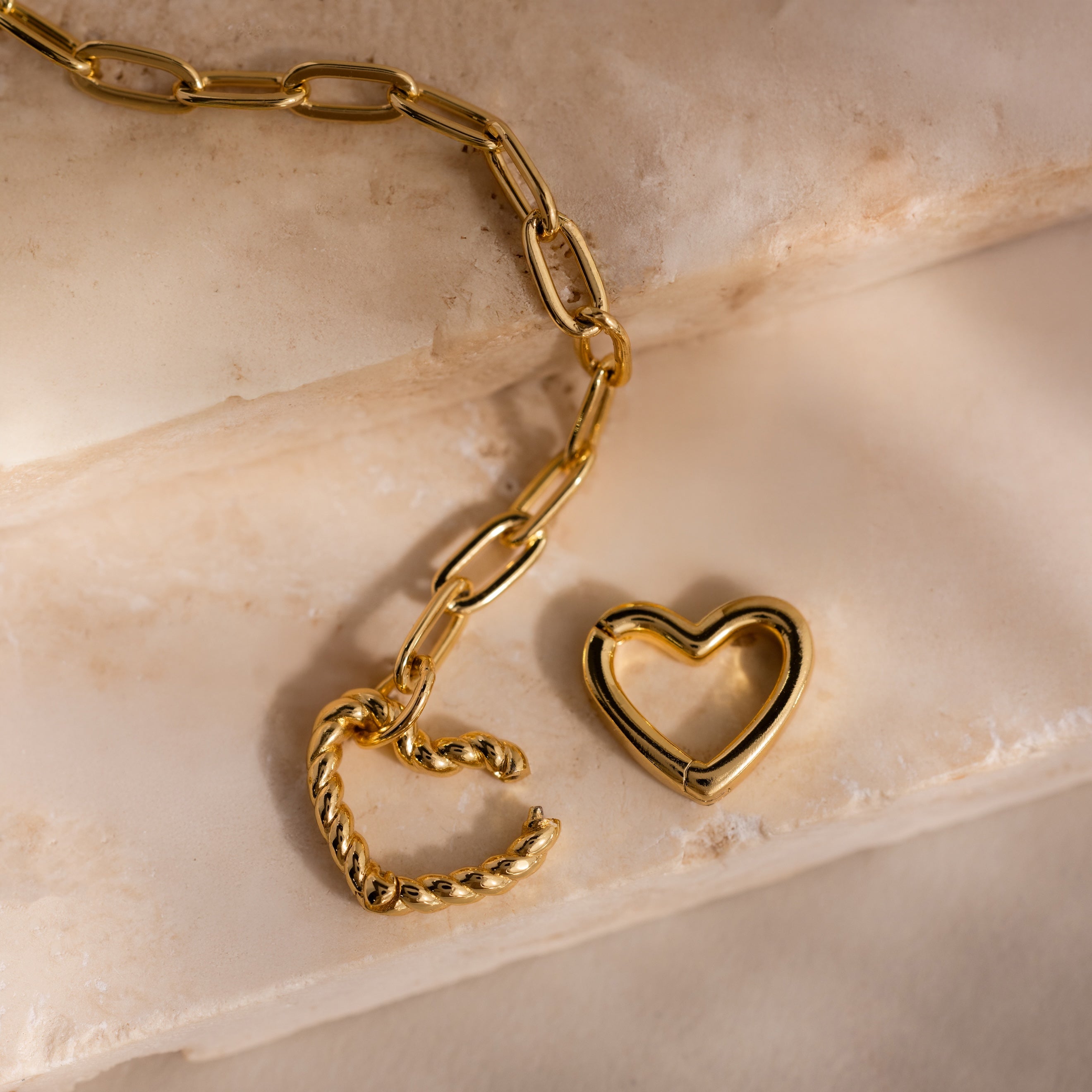 Gold Heart Carabiner Bracelet with paperclip chain links and two heart-shaped clasps—one twisted and one smooth—resting on stone steps.